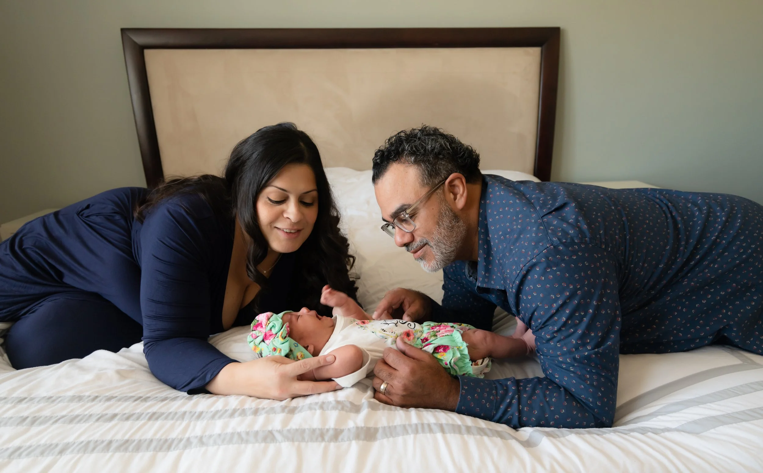 A woman and a man with glasses and gray hair are lying on a bed, looking at a newborn baby girl who is lying between them. The woman has long dark hair and is wearing a dark blue dress. The man is wearing a blue shirt with a pattern.