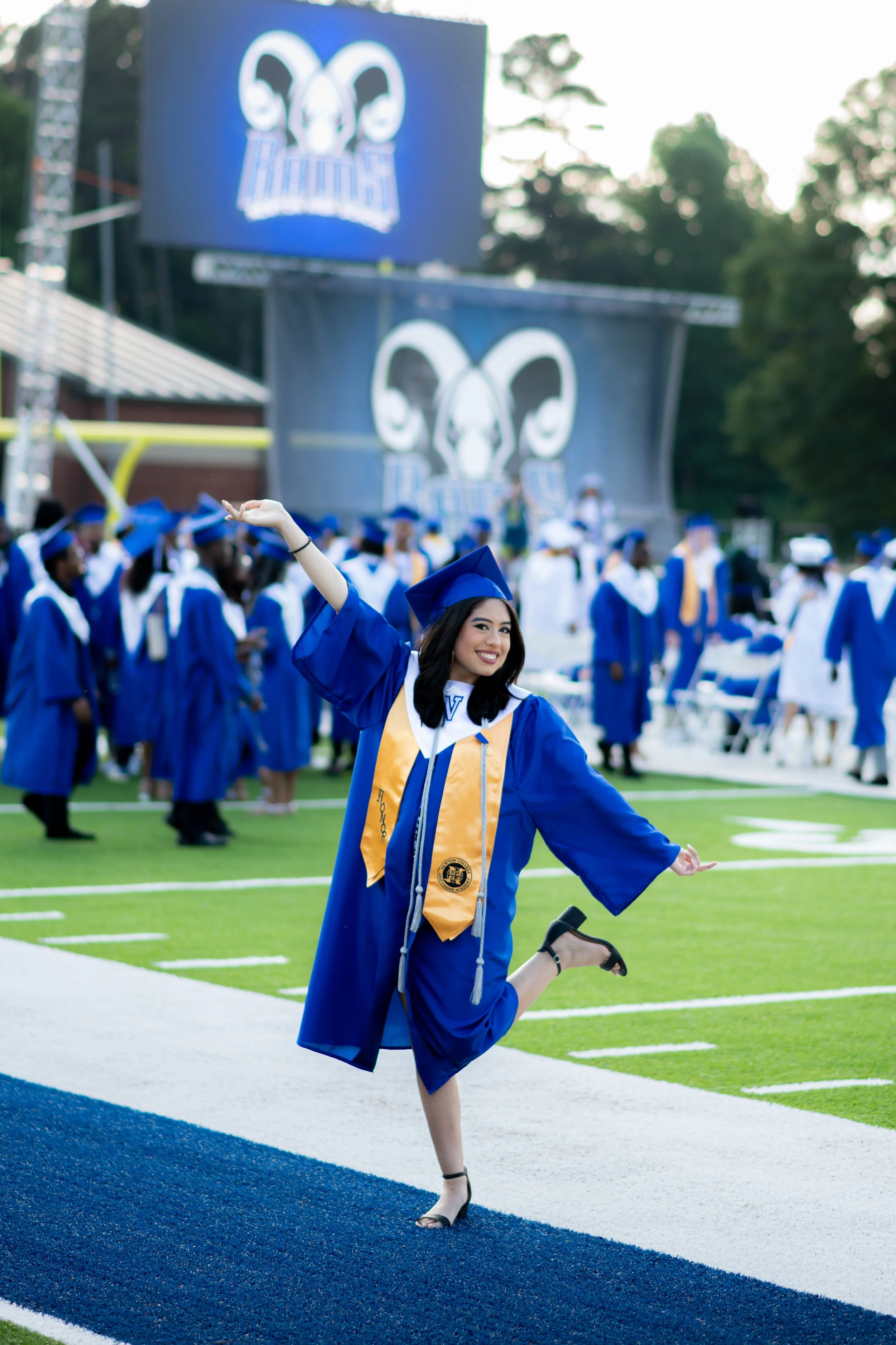 A smiling graduate in a blue cap and gown with a yellow stole celebrating on a football field during a graduation ceremony, with other graduates in the background.