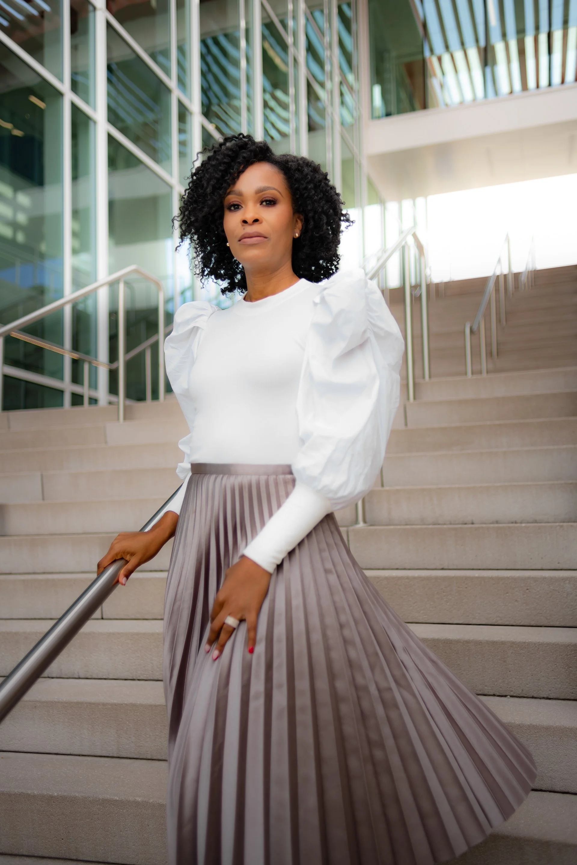 A woman with curly black hair wearing a white blouse with puffed sleeves and a pleated taupe skirt standing on stairs outside a modern glass building.