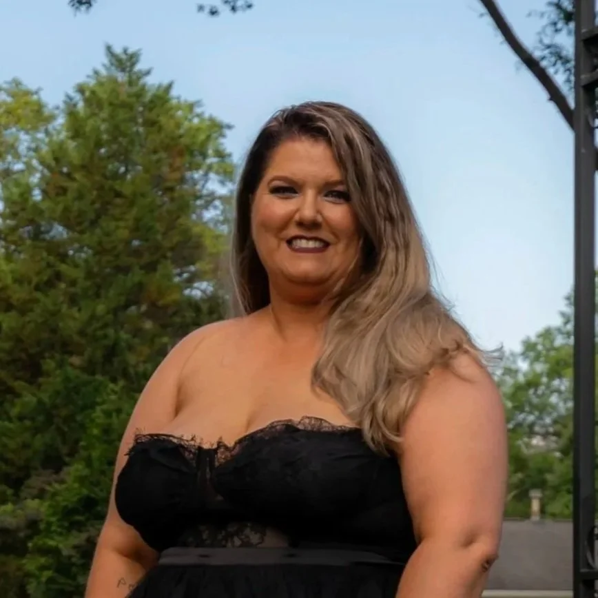 A woman with long, wavy hair smiling outdoors, wearing a black strapless top with lace details, surrounded by trees and a clear sky.