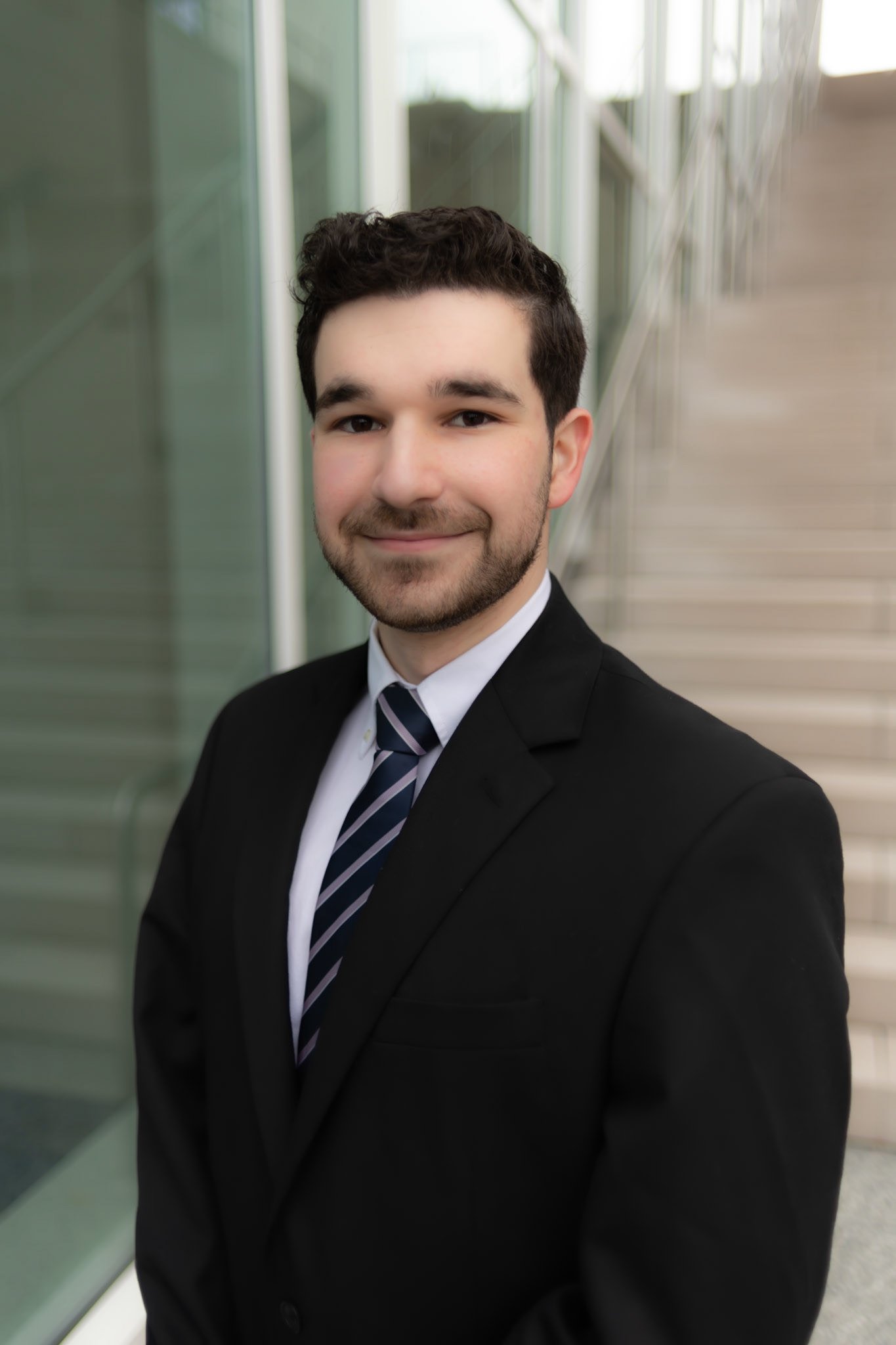A young man with dark, curly hair and a trimmed beard, dressed in a black suit, white shirt, and a striped tie, stands in front of a modern building with glass windows and concrete stairs.