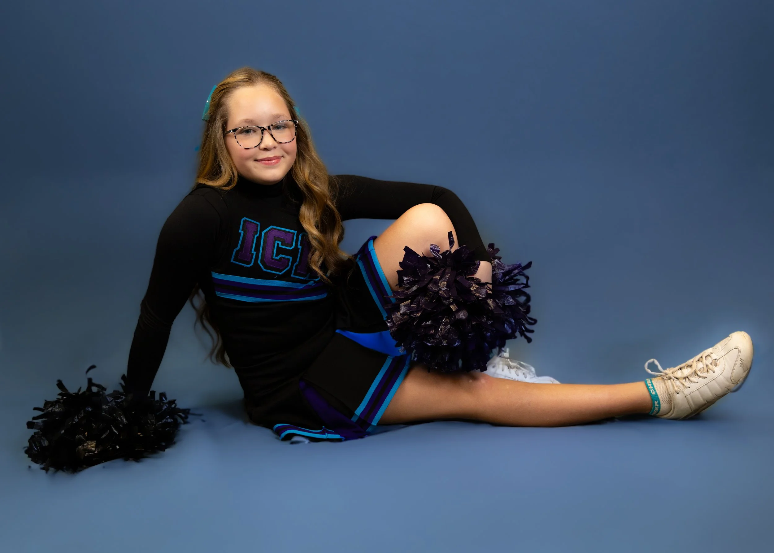 A young girl wearing glasses, cheerleading uniform with 'ICE' on it, sitting on a blue studio background. She holds black pom-poms, with one resting on her lap and the other on the floor. She is smiling, with long wavy hair, and is wearing white snea