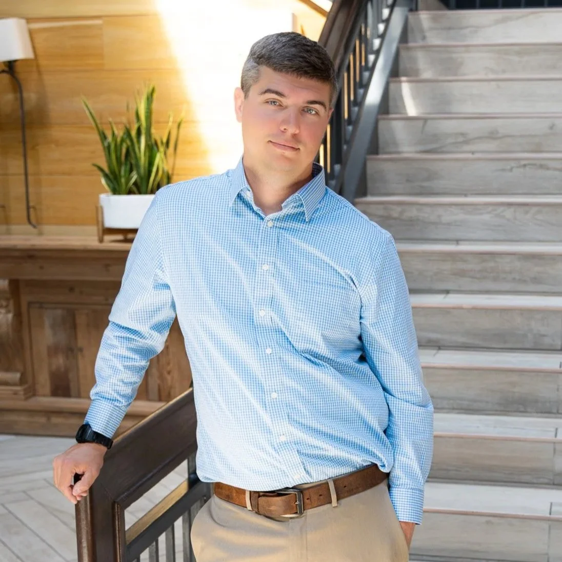 A young man, Austin, with short dark hair, wearing a light blue checkered shirt and beige pants, leaning on a wooden railing inside a modern building with wooden accents and a staircase in the background.