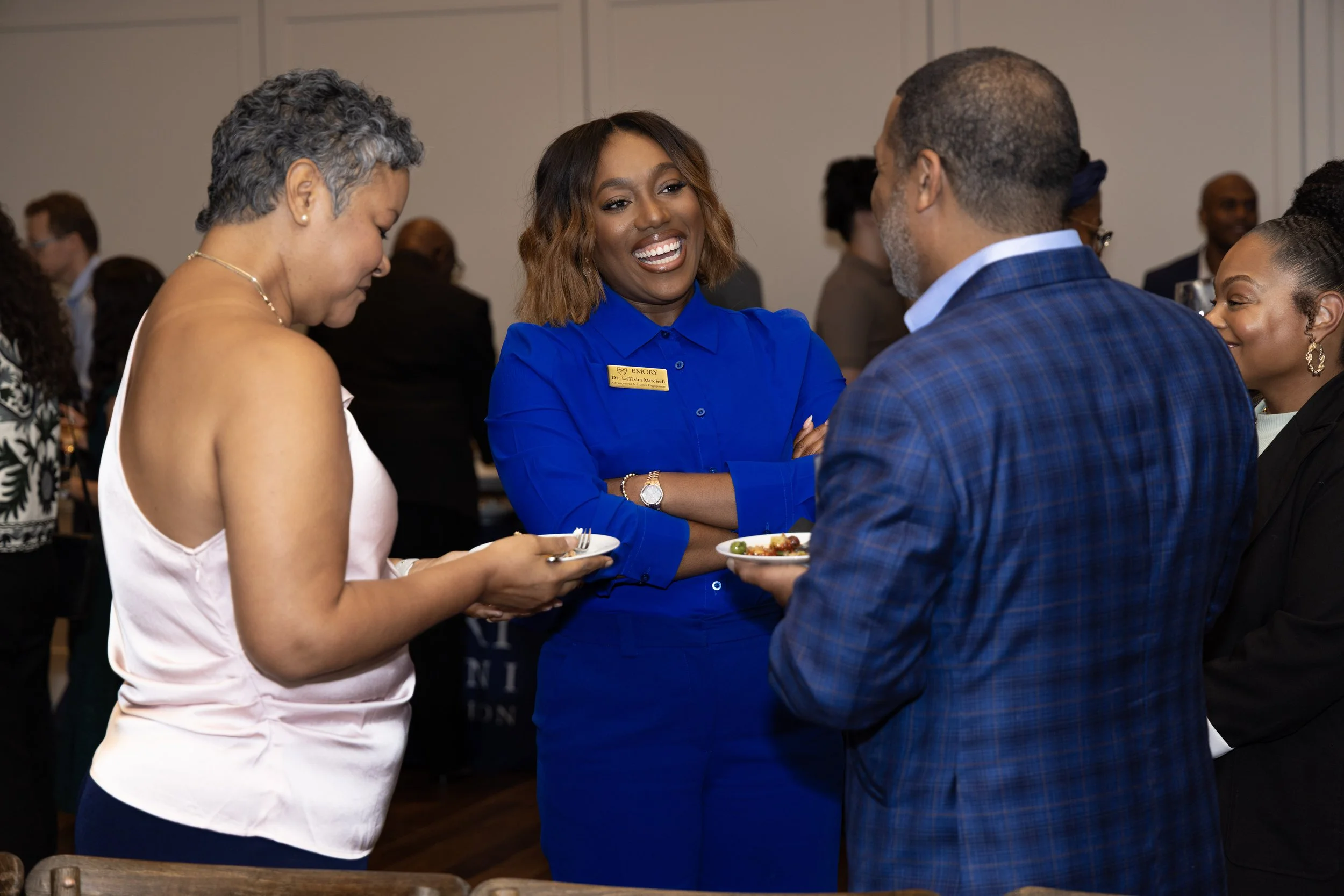 Group of four diverse people in a conversation at a formal event, with women and men, holding plates with small portions of food, indoors with a plain background.