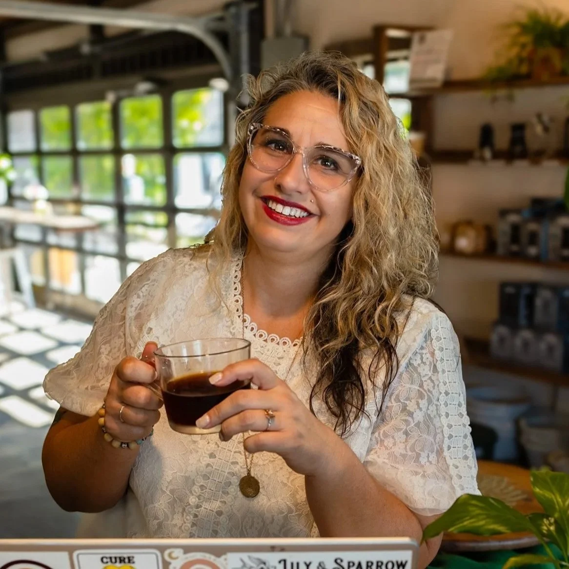 A woman, Noelle Cosby, with curly blonde hair, wearing glasses and a white lace top, smiling while holding a cup of coffee in a cozy cafe with large windows and wooden shelves in the background at Lily and Sparrow Mercantile.