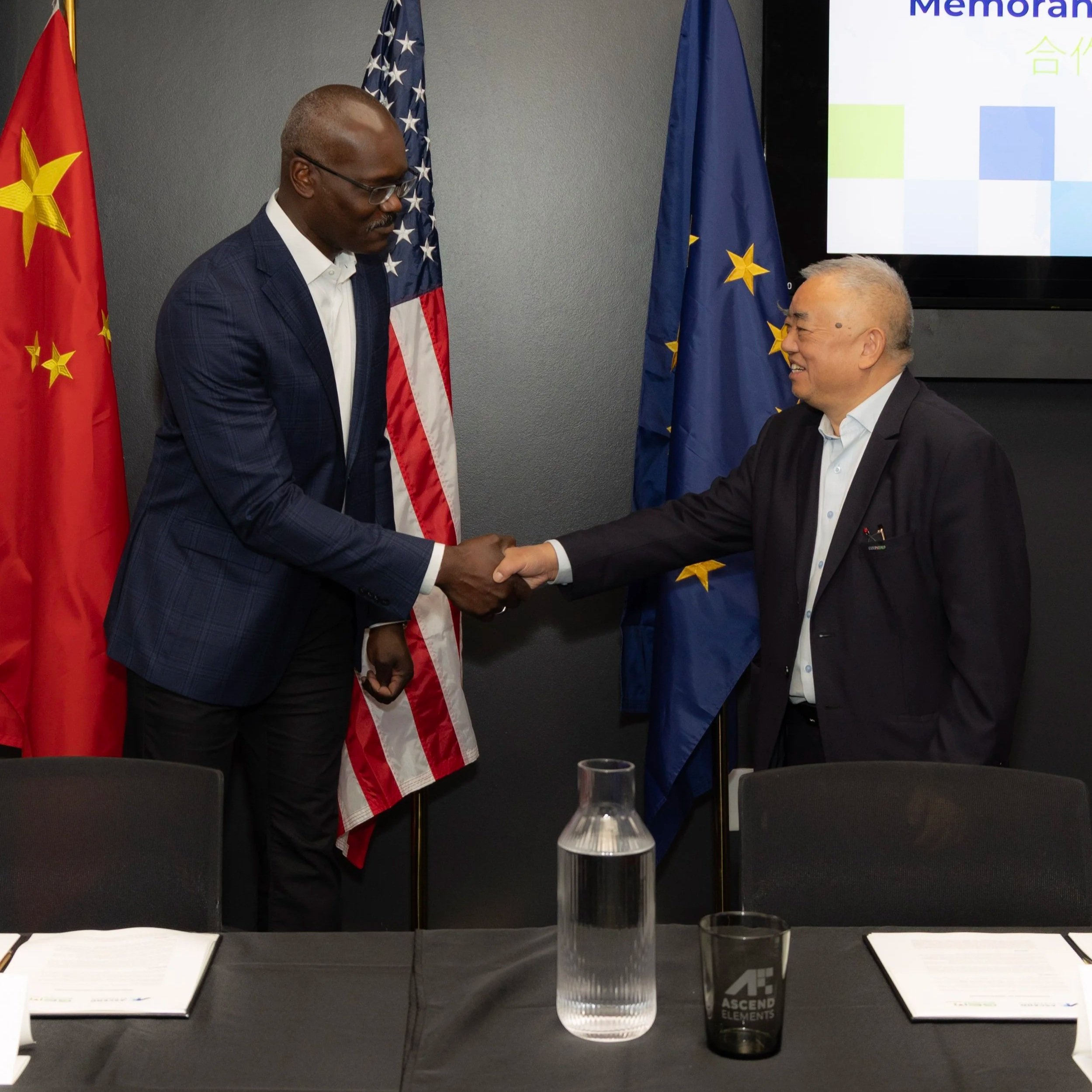 Two men shaking hands in a meeting room with flags of China, the US, and the European Union in the background, and a table with documents, a water pitcher, and a glass in the foreground.