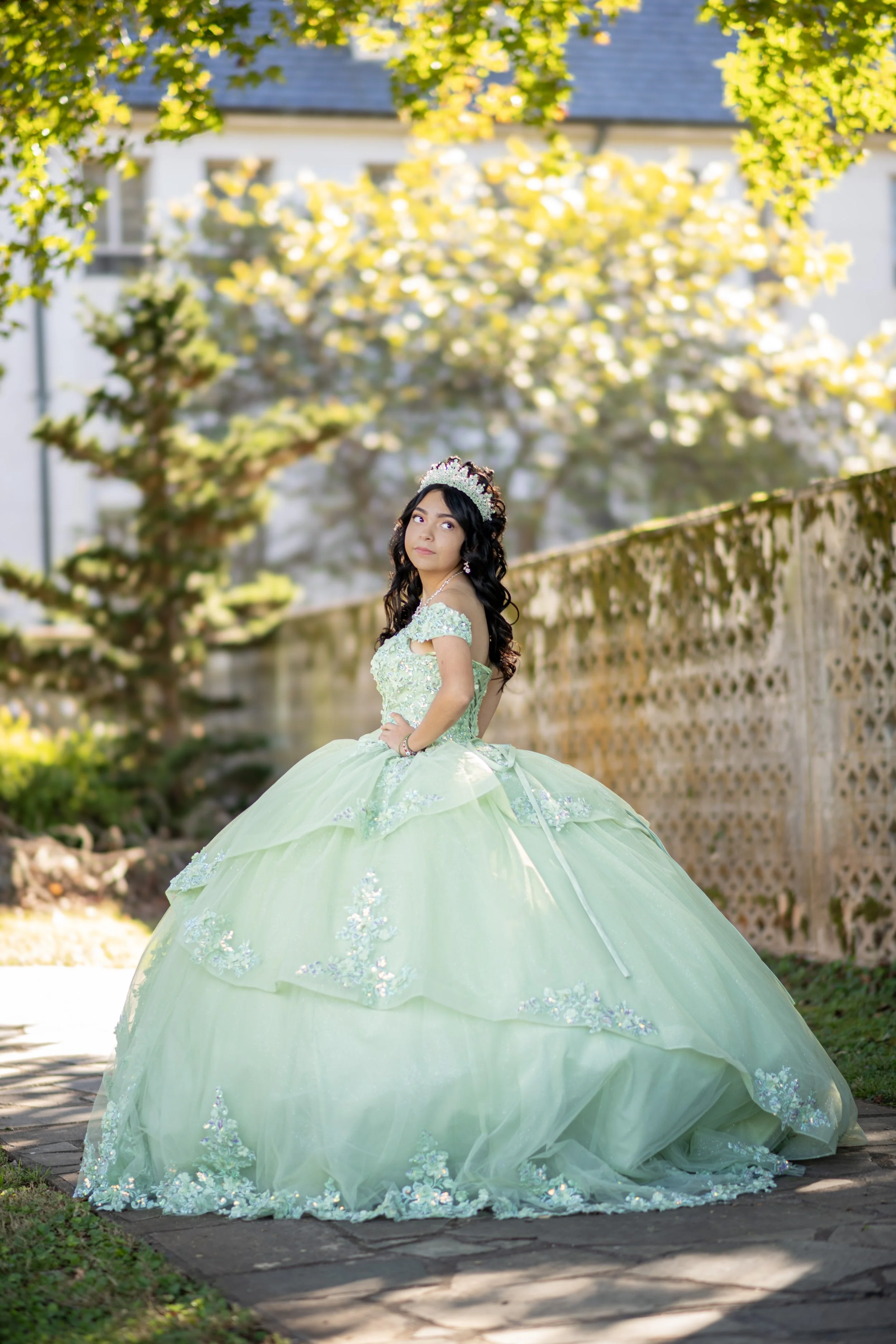A girl dressed as a princess in a light green ball gown with floral embellishments, wearing a tiara, standing outdoors on a stone path, with trees and a fence in the background.