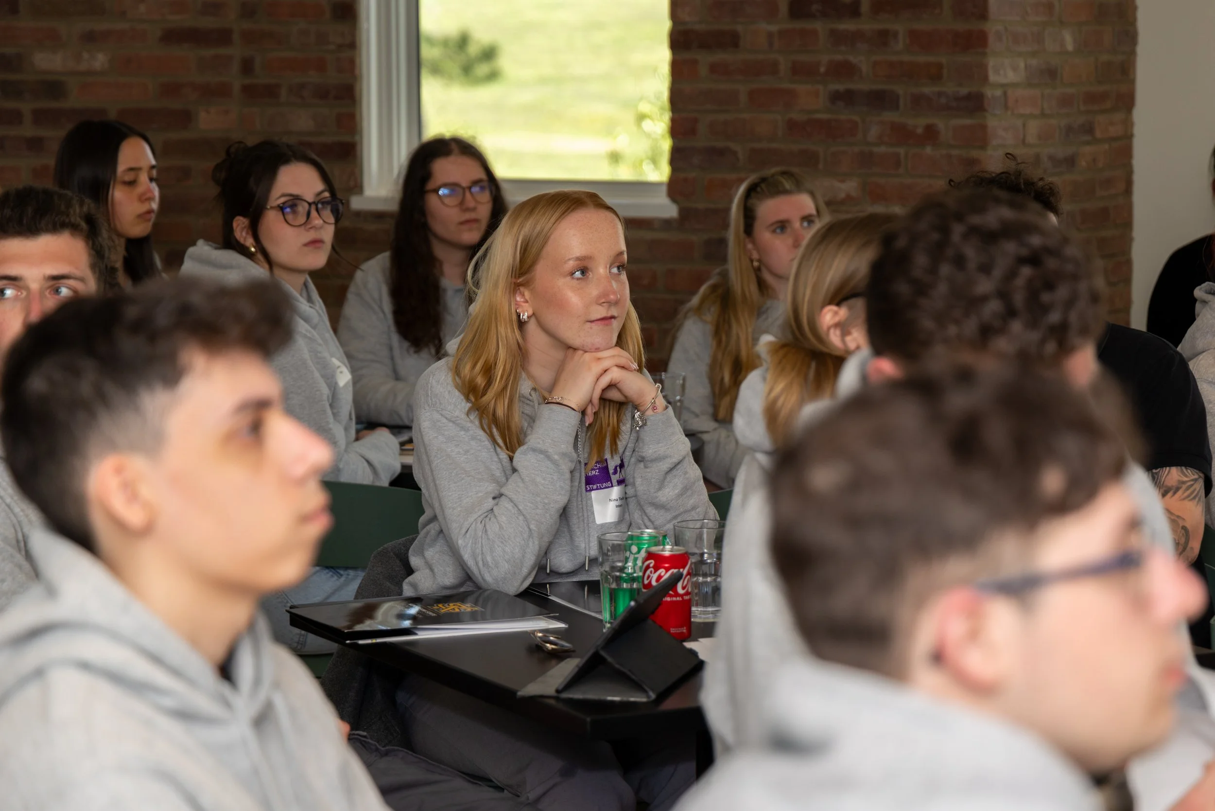 Young adults attending a lecture or seminar in a classroom setting, wearing grey hoodies, sitting at tables with drinks and notebooks, with brick walls and a window in the background.