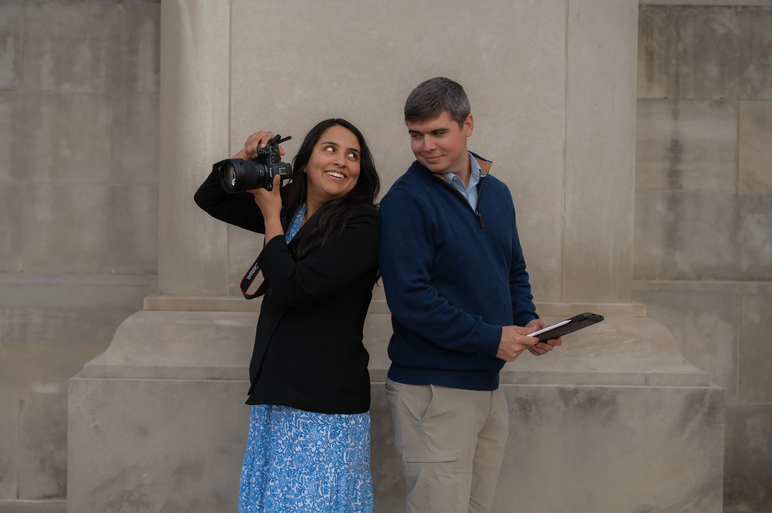 A woman holding a camera on her shoulder and smiling at a man, who is holding a tablet, against a stone wall background.