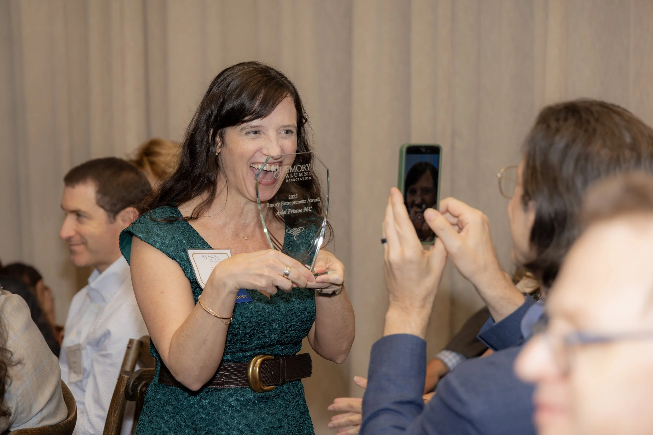A woman in a teal dress holding a glass award, smiling, while someone takes her photo at an event.