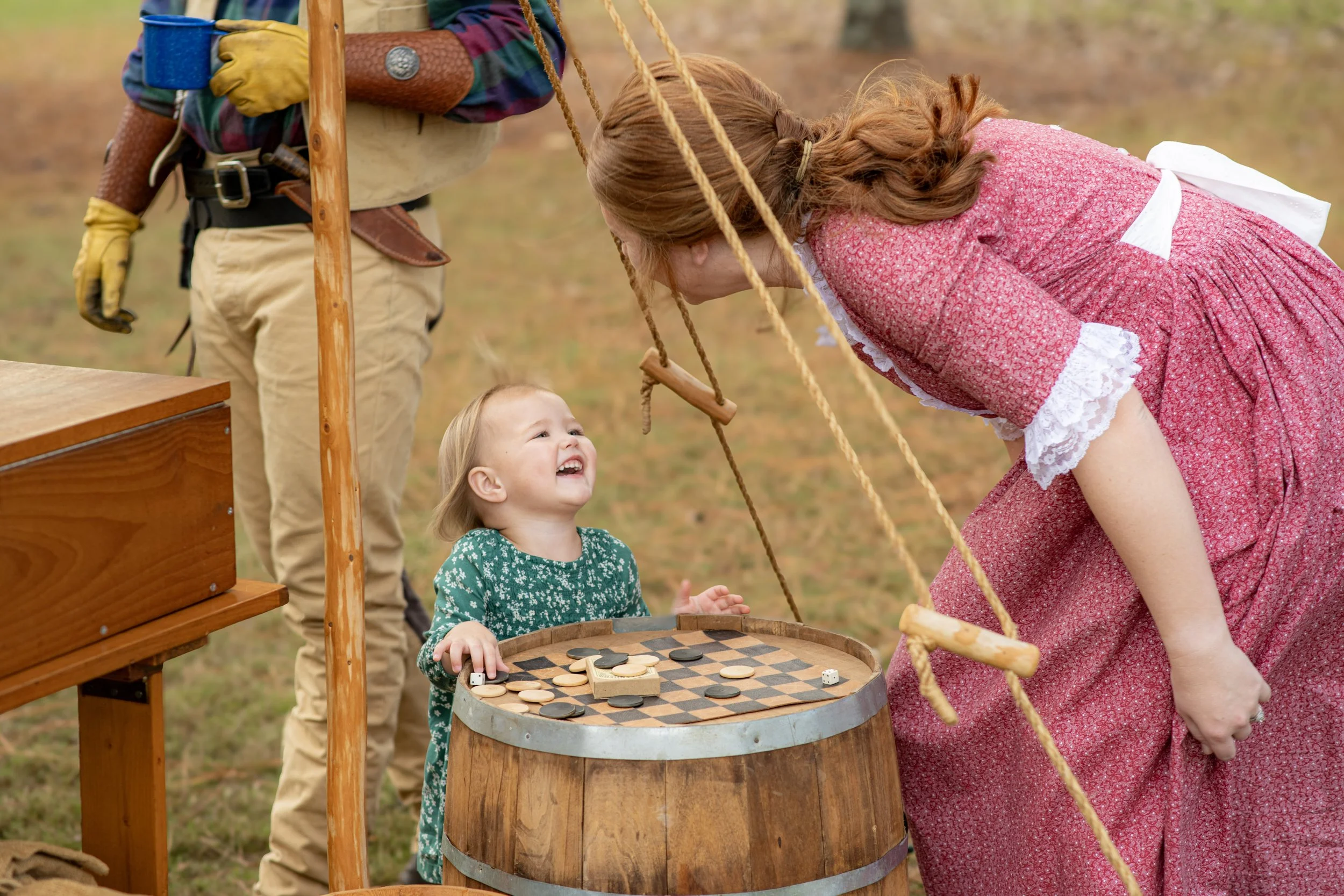 A young girl and a woman bent over a barrel playing checkers outdoors with a woman dressed in period clothing. A man in period attire stands nearby. The scene appears historical or reenactment.