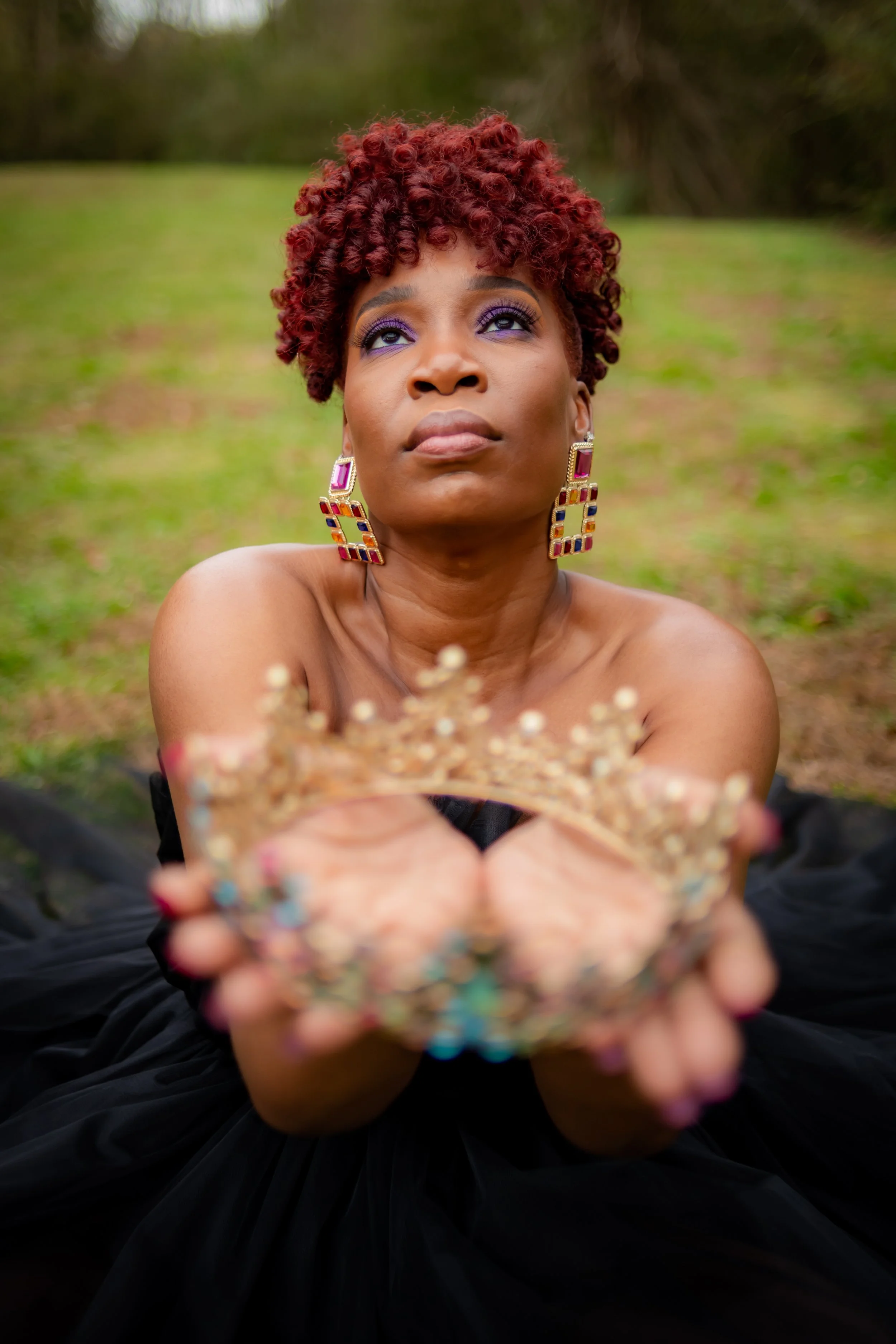 A woman with short, curly red hair and colorful earrings holding a jeweled crown outdoors, looking up with a contemplative expression.