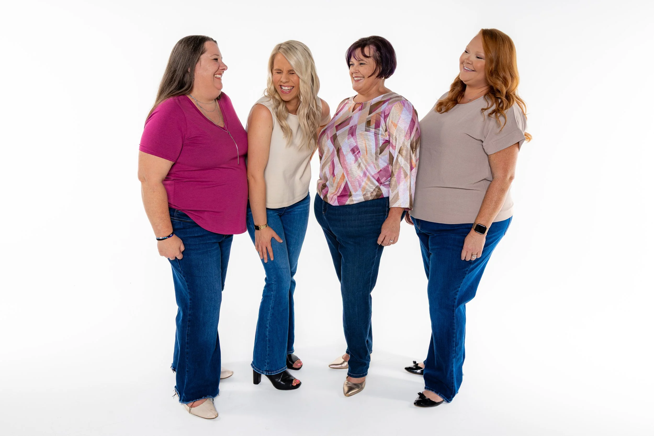 Four women with different hair colors standing together, smiling and laughing in a photo studio with a white background.
