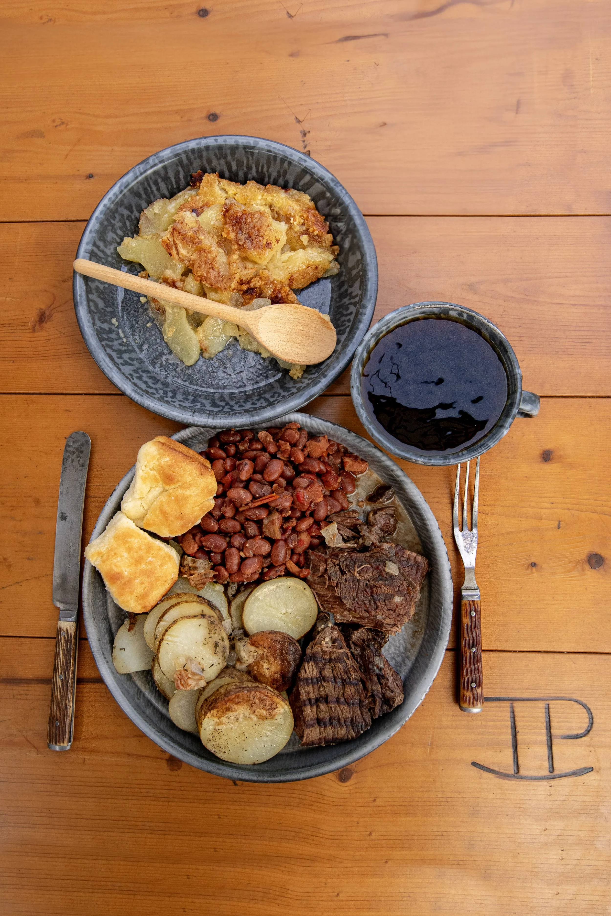 A plate of barbecue food with baked potatoes, baked beans, and brisket, along with a biscuit, a cup of dark soda, and a smaller plate of potato casserole on a wooden table with a fork and knife.