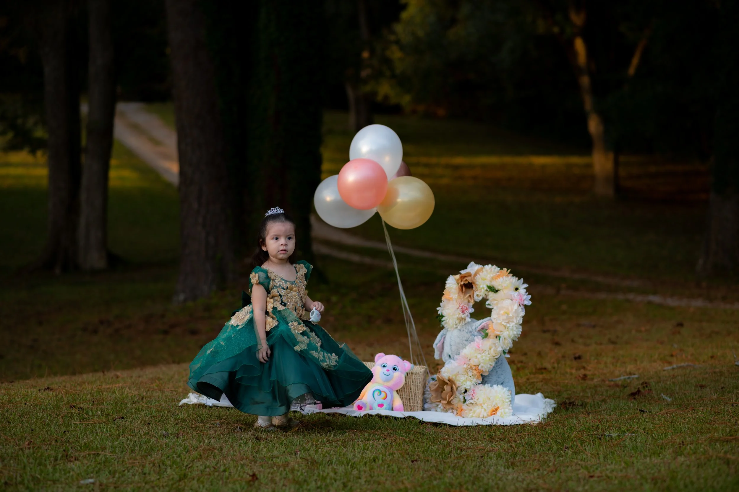 Young girl in a dark green and gold dress standing outdoors near balloons and a floral number two decoration, with plush toys and a wreath in the background, during evening.