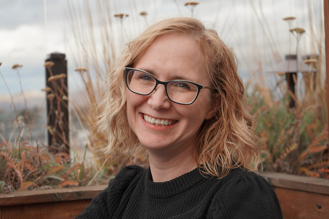 A smiling woman with short, curly blonde hair and glasses, sitting outdoors near a wooden railing with grasses and a cloudy sky in the background.