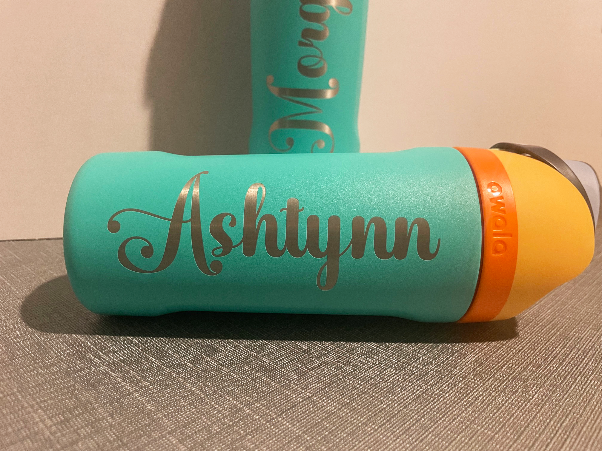 A teal-colored water bottle with the name "Ashtynn" written on it in cursive, placed on a textured surface with another similar bottle in the background.