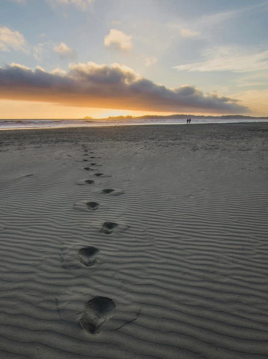 Footprints in the sand on a beach leading towards the horizon, with two people walking in the distance during sunset or sunrise, with clouds and calm ocean waves visible.