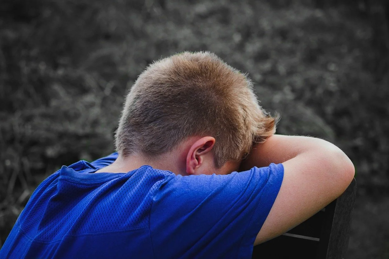 A young boy with blonde hair and wearing a blue shirt, sitting outdoors with his head resting on his bent arm, appearing to be upset or deep in thought, against a blurred natural background.