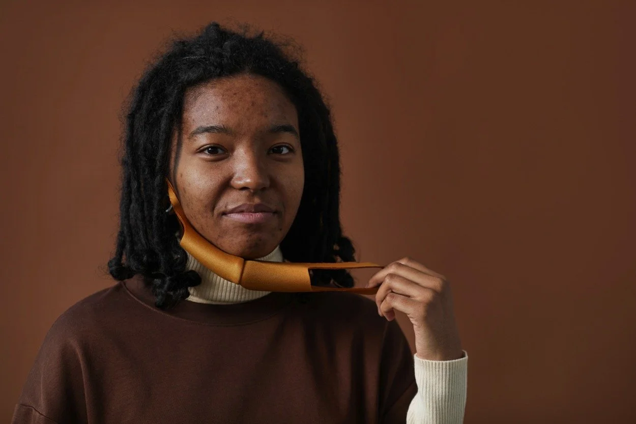 Young woman with dreadlocks holding a gold telephone receiver to her neck while looking at the camera, against a brown background.