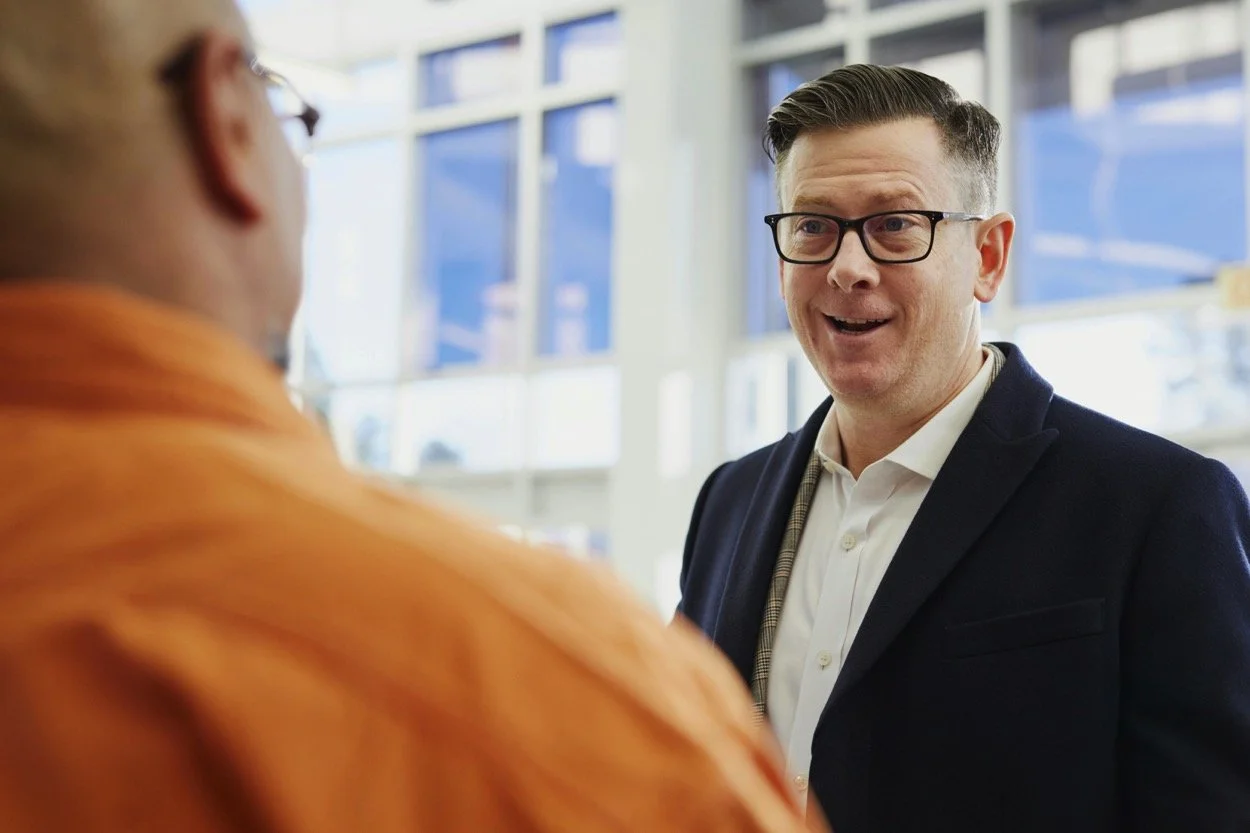 A man with glasses and a suit talking to another person in an indoor setting with large windows.