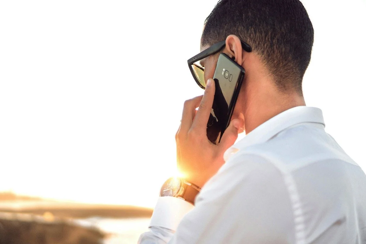 A man wearing sunglasses and a white shirt talking on a smartphone, with a sunset in the background.