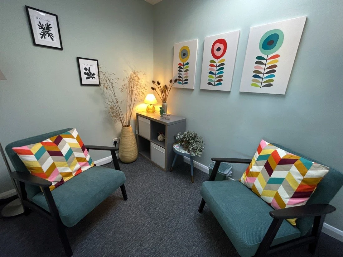 A cozy waiting room with two teal armchairs, each with a colorful geometric patterned pillow. A small gray shelf with decorative items and a lit lamp is in the corner. The wall has colorful abstract flower artwork, and there are two black and white framed botanical prints on the left wall.