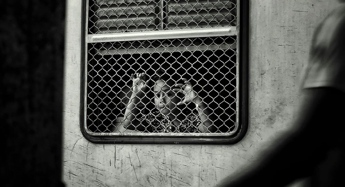 A woman looking through a barred window with a serious expression, her hands pressed against the bars.