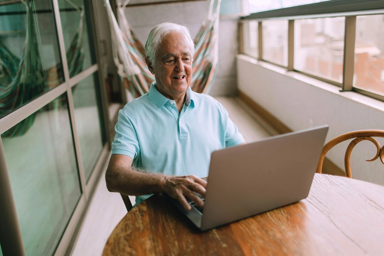 An elderly man with white hair sitting at a wooden table, smiling and using a laptop, on a balcony with large windows and a view of neighboring buildings.