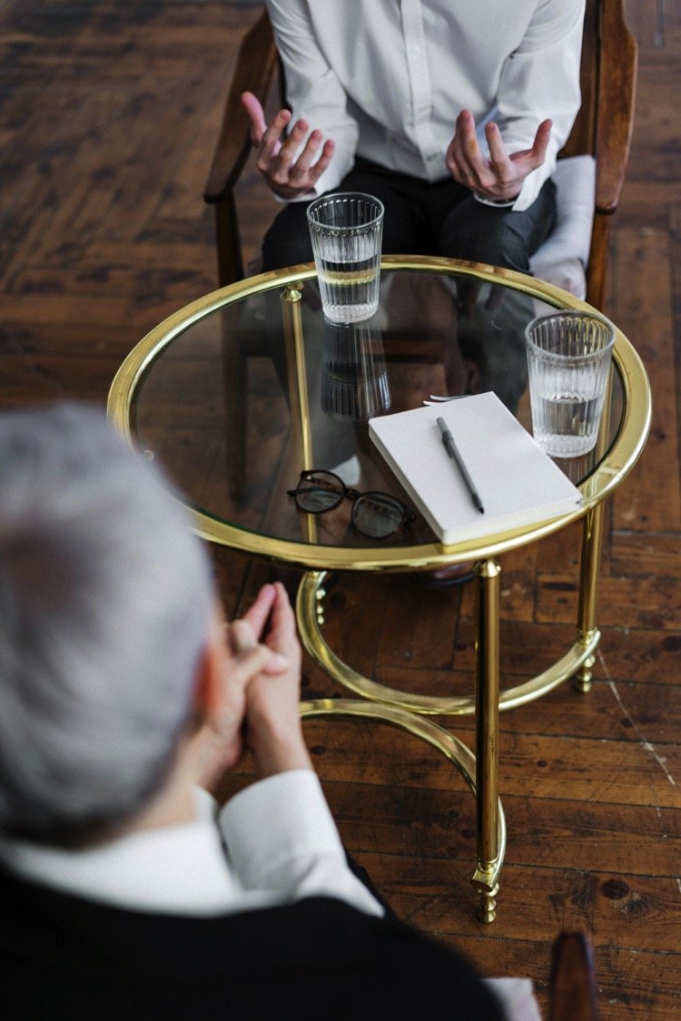 Two people having a conversation at a glass-top table with two glasses of water, a notebook, a pen, and eyeglasses; one person with white hair and black clothing, the other with dark hair and a white shirt, seated in a room with wooden flooring.