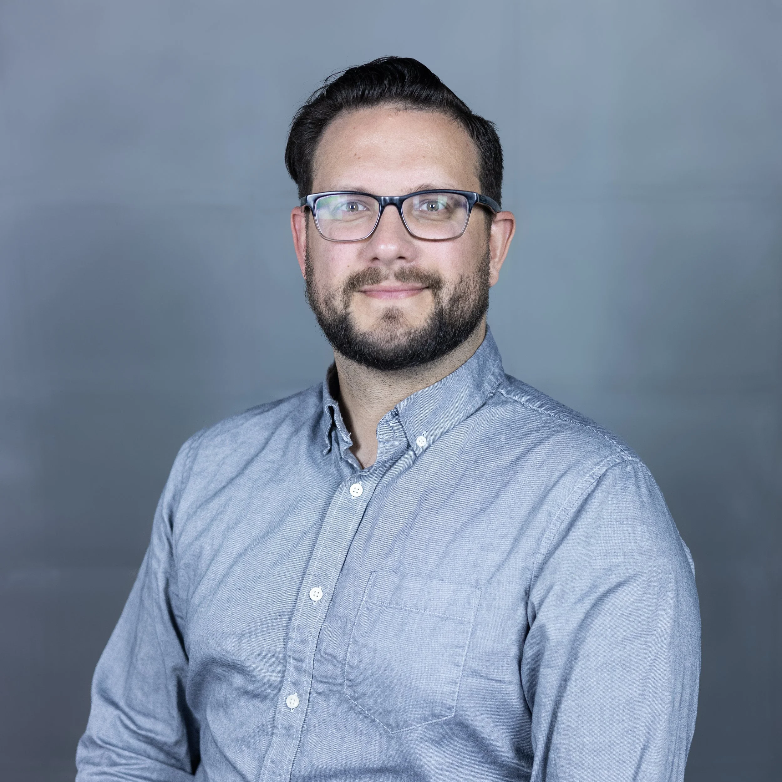A man with dark hair, glasses, and a beard, wearing a light blue button-up shirt, smiling at the camera against a plain gray background.