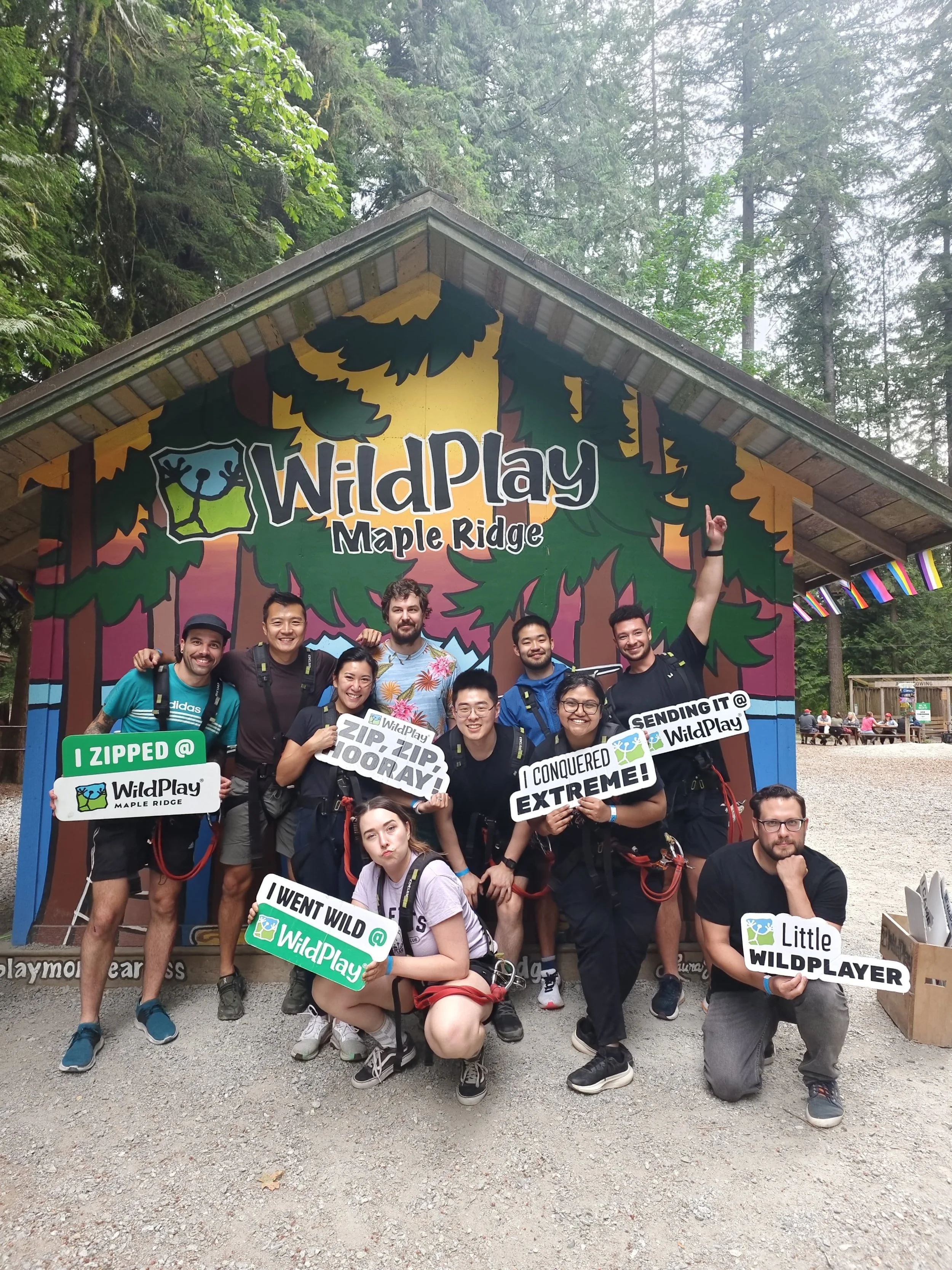 Group of nine people posing in front of a sign for WildPlay Maple Ridge, holding various signs and making celebratory gestures during an outdoor adventure activity in a forest setting.