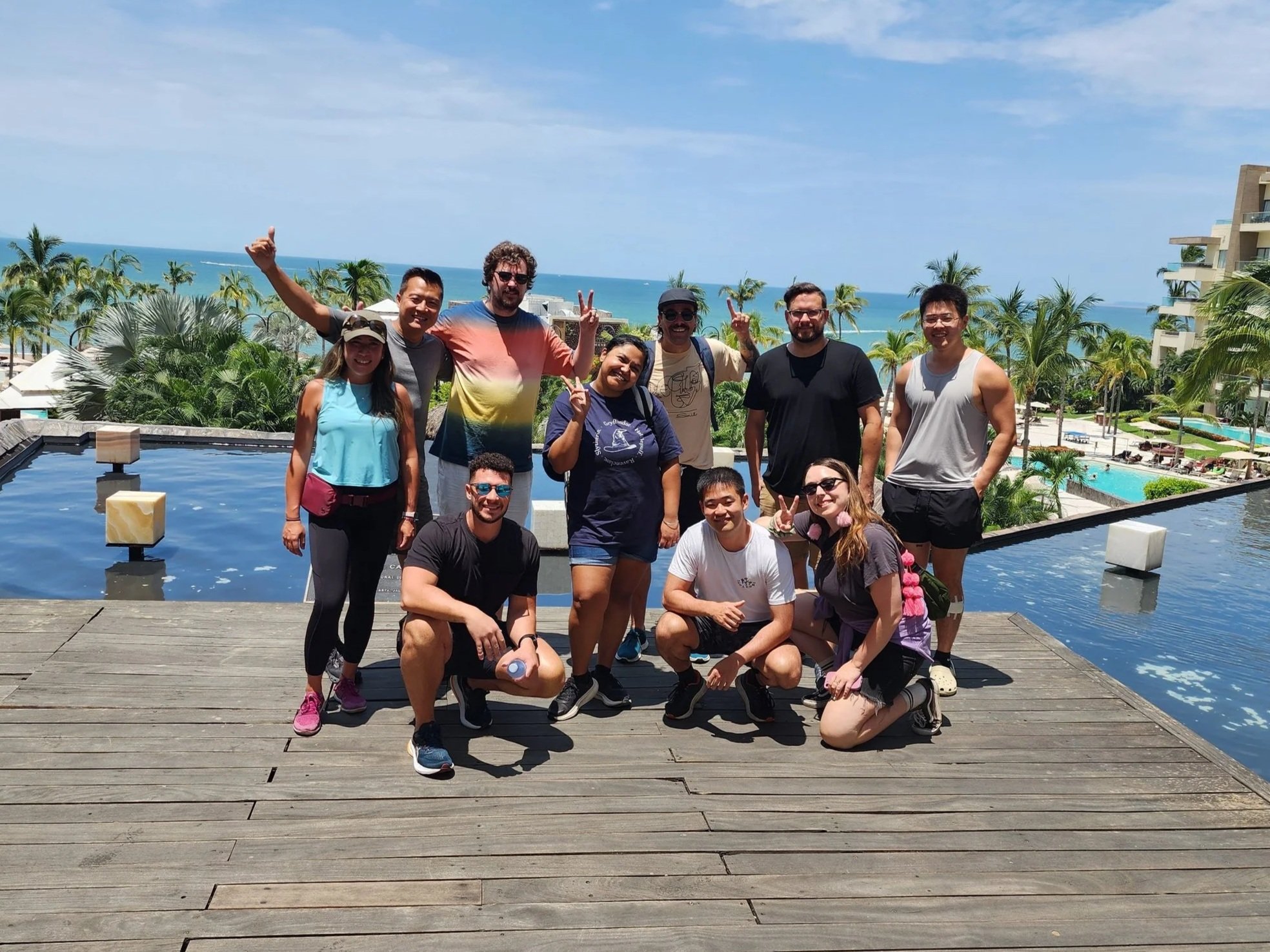 Group of eleven people posing together on a wooden deck at a hotel with an ocean view, palm trees, and swimming pools in the background, during a sunny day.