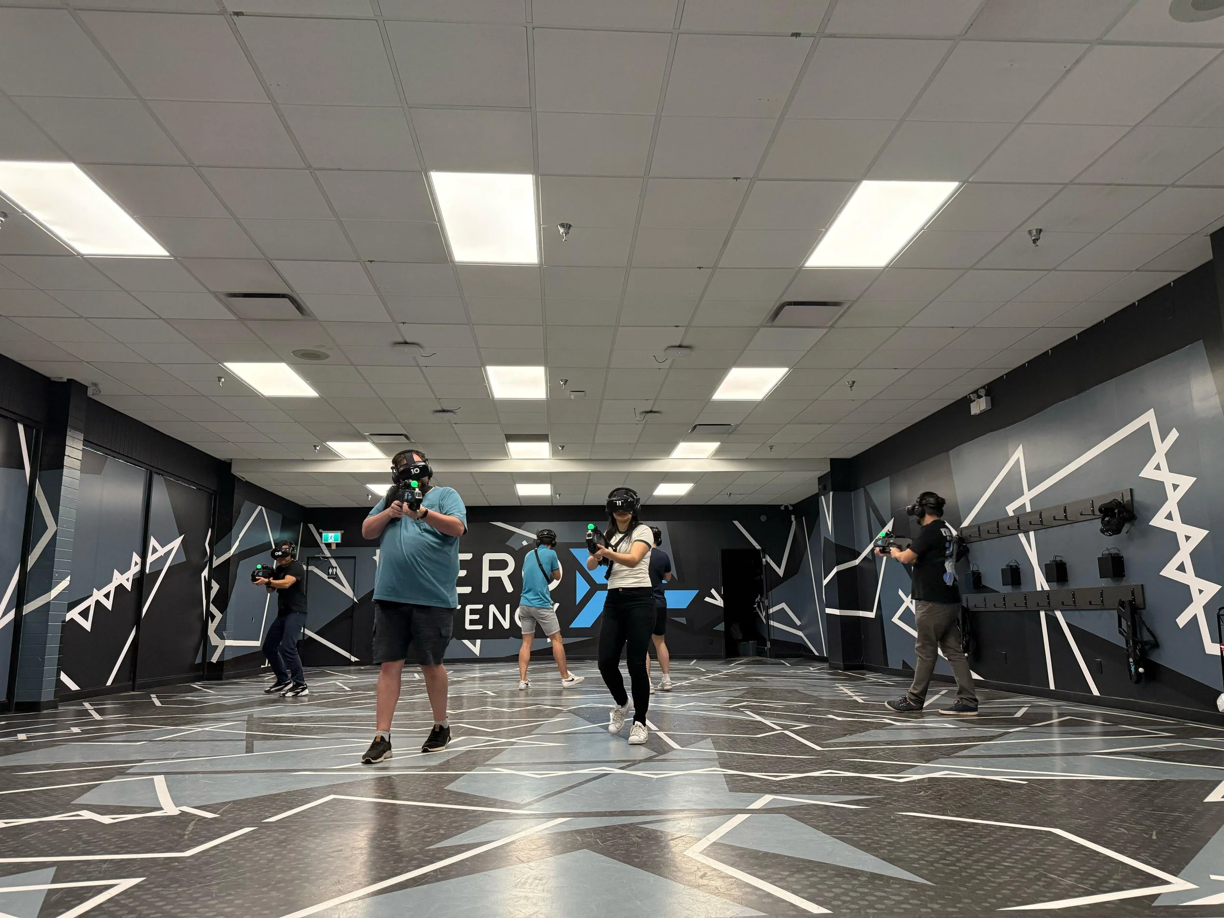 People wearing virtual reality headsets and holding controllers in a large indoor space with black and gray geometric wall patterns.