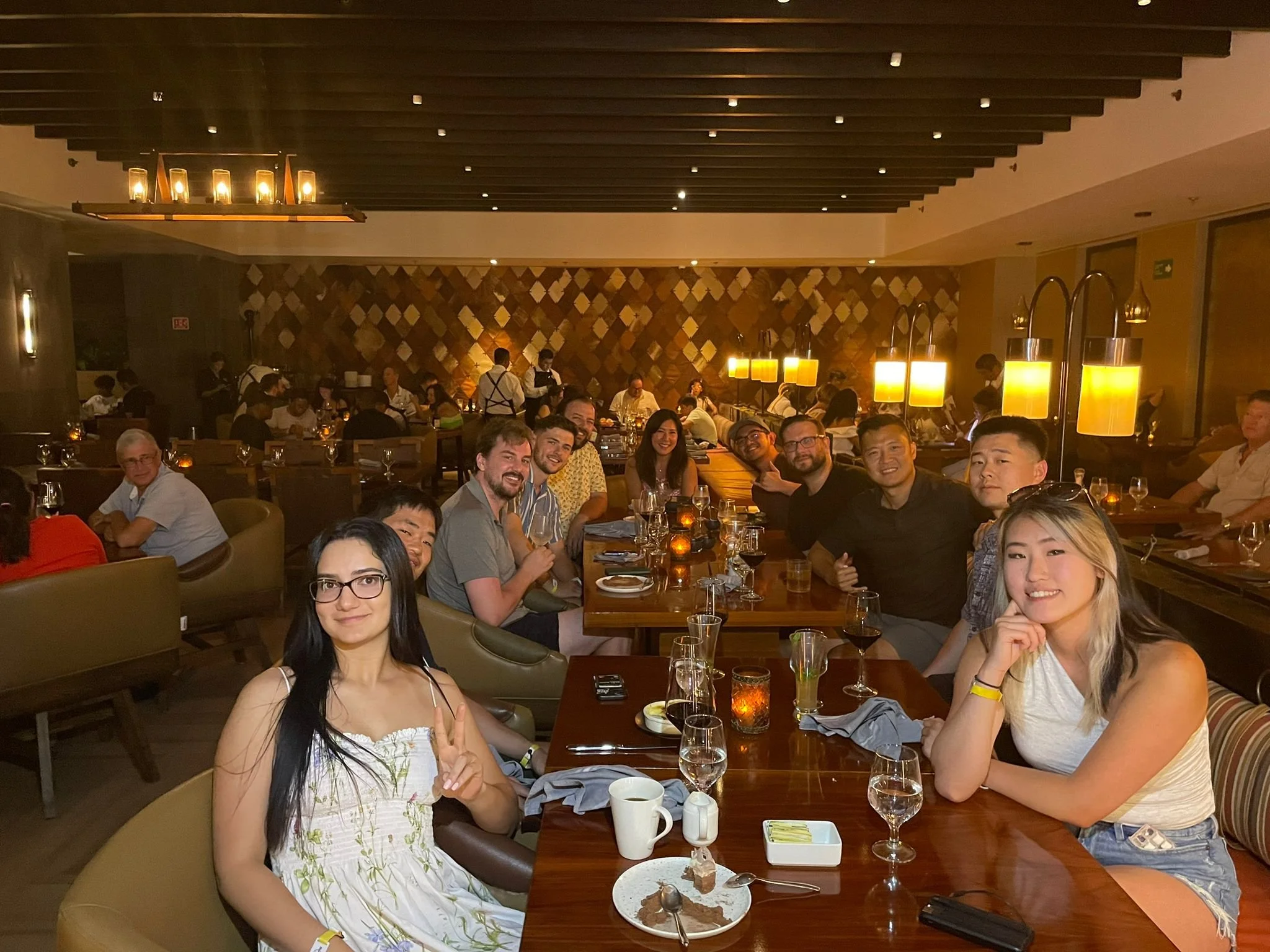 Group of people enjoying dinner at a restaurant, sitting at a long table with glasses, cups, and plates, with a warm, dimly-lit ambiance and wooden decor.
