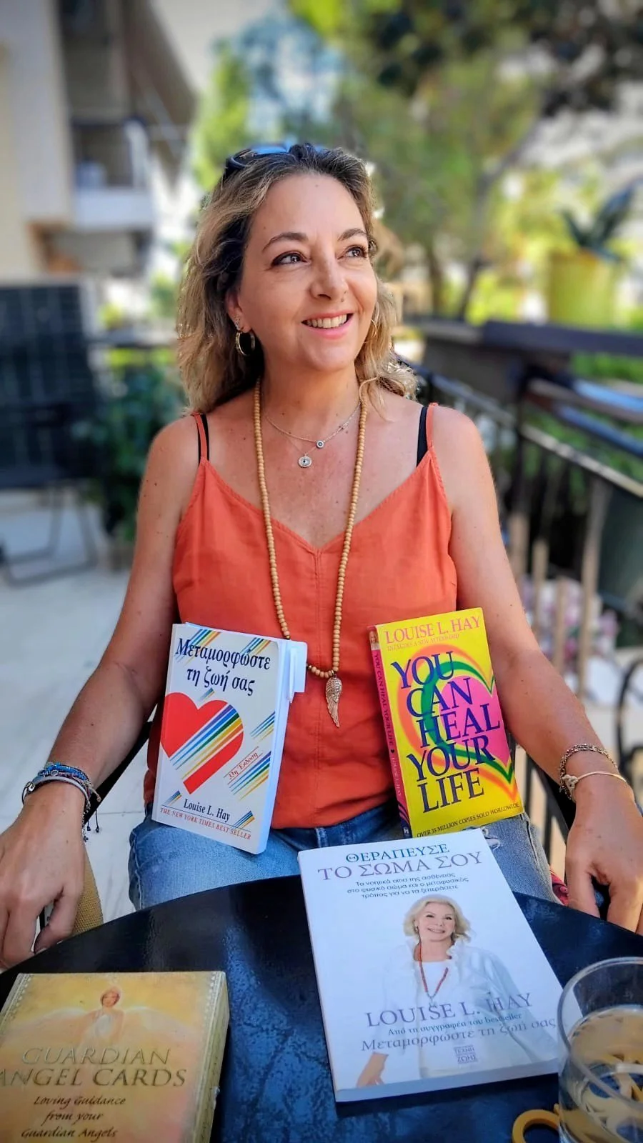 A woman with wavy blonde hair wearing an orange tank top, layered necklaces, and bracelets, sitting at a table outdoors with books about healing and spirituality, smiling and looking away.