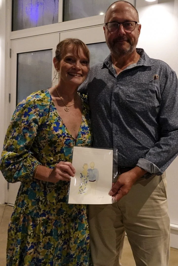A smiling woman in a floral dress holding an artwork of a man and woman, standing next to a man in a patterned shirt, inside a room with large windows.