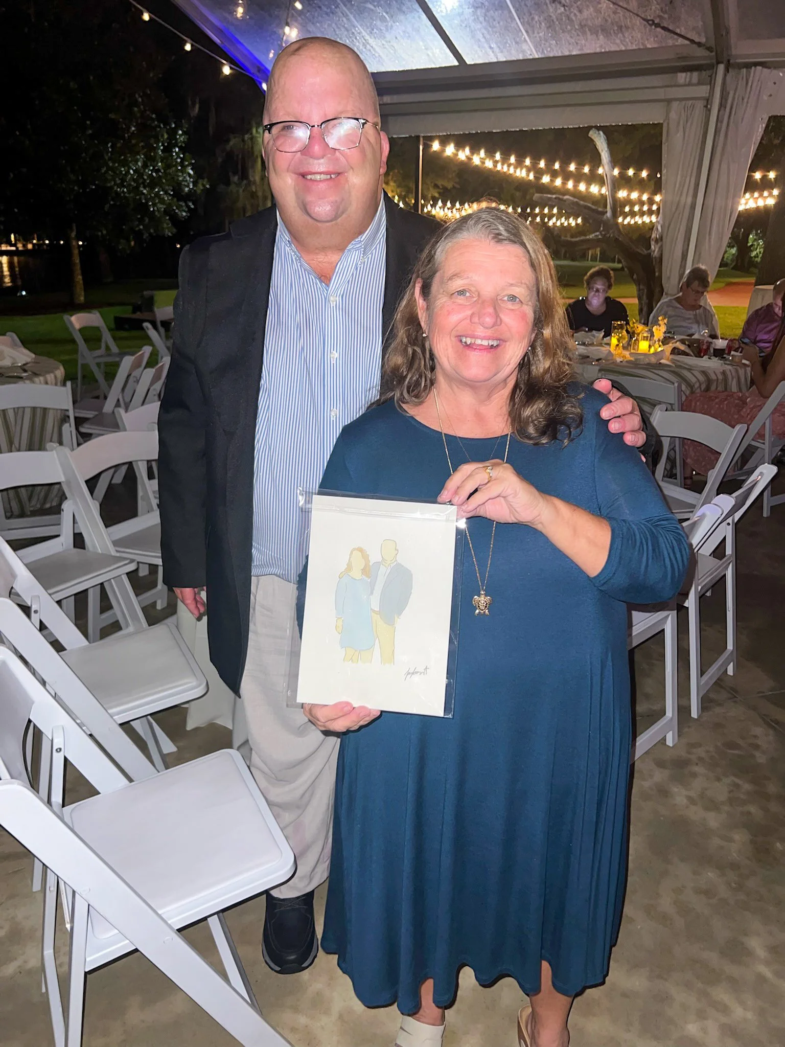 A man and woman smiling at a social event under a tent, with the woman holding a card with a drawing of a couple on it, surrounded by string lights.