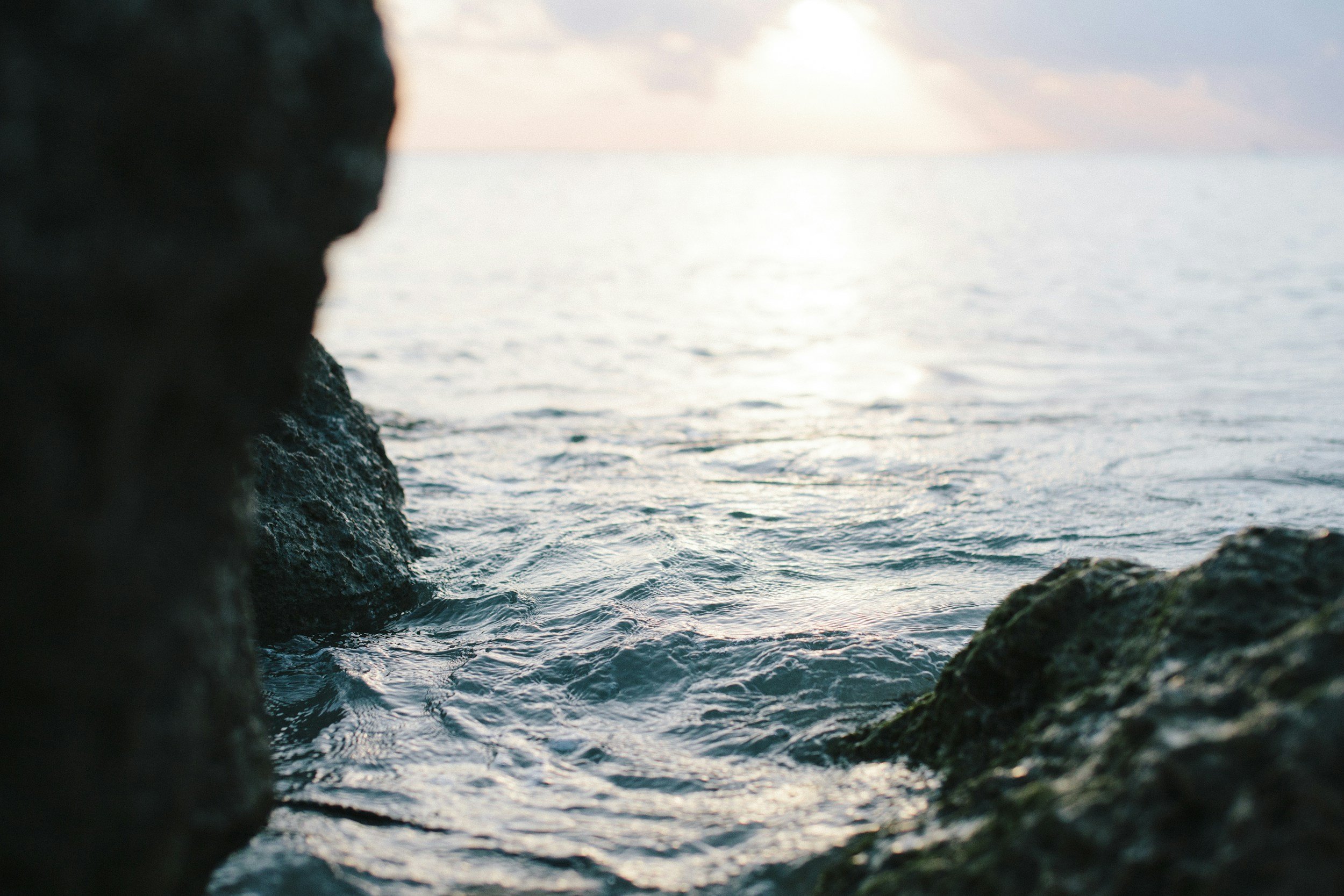 View of ocean water with rocks in the foreground, sunset sky in the background.