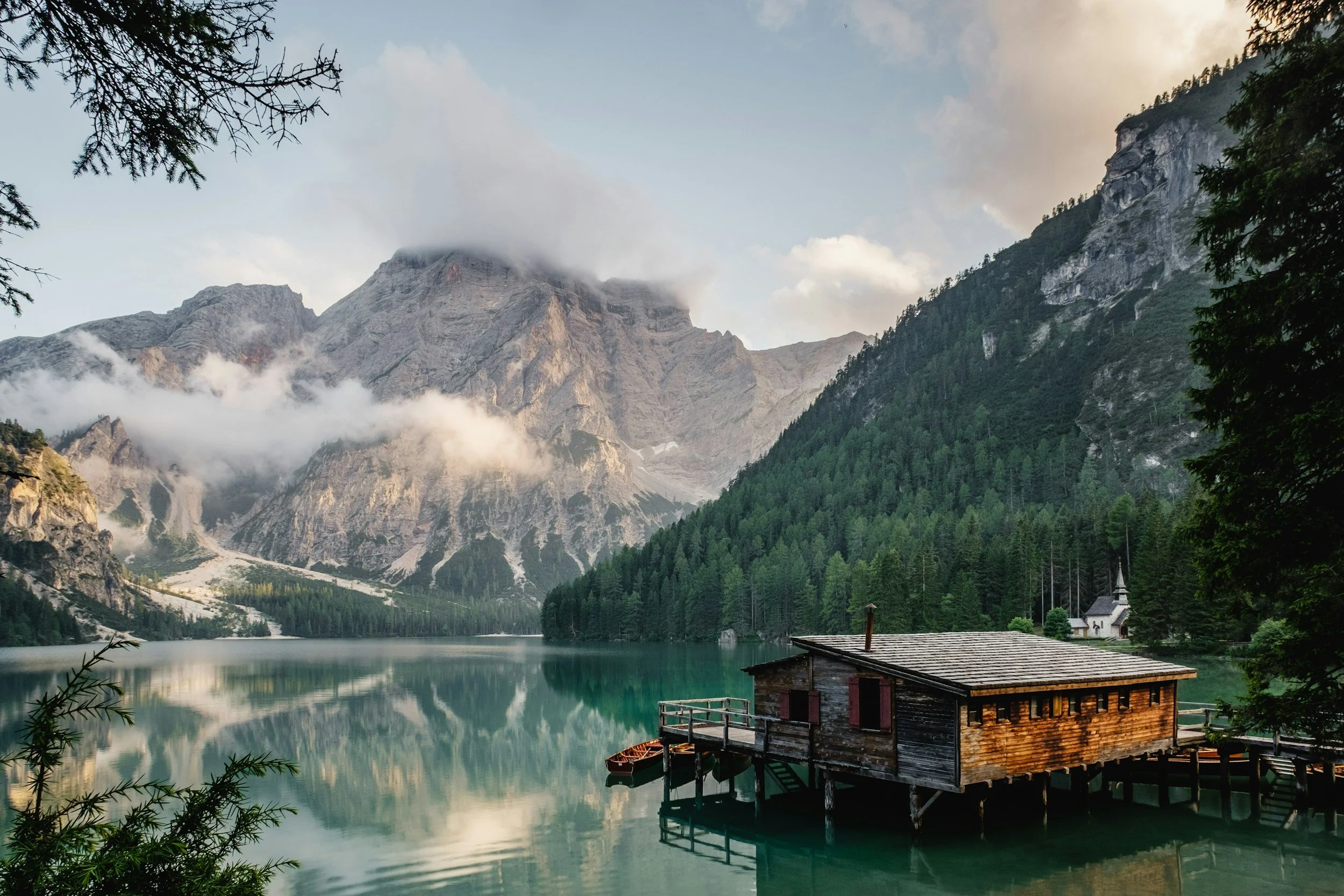 A wooden boathouse on stilts in a calm mountain lake surrounded by pine trees and steep rocky mountains with some clouds overhead.