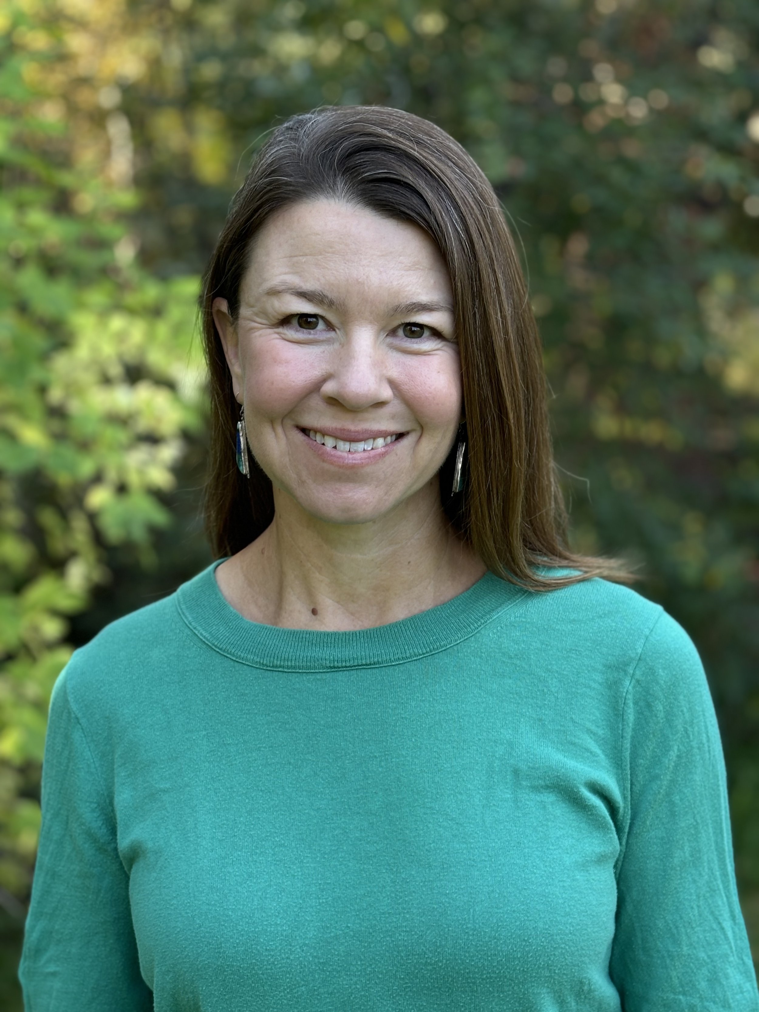 A woman with long brown hair wearing a turquoise shirt and earrings, smiling outdoors with green foliage in the background.