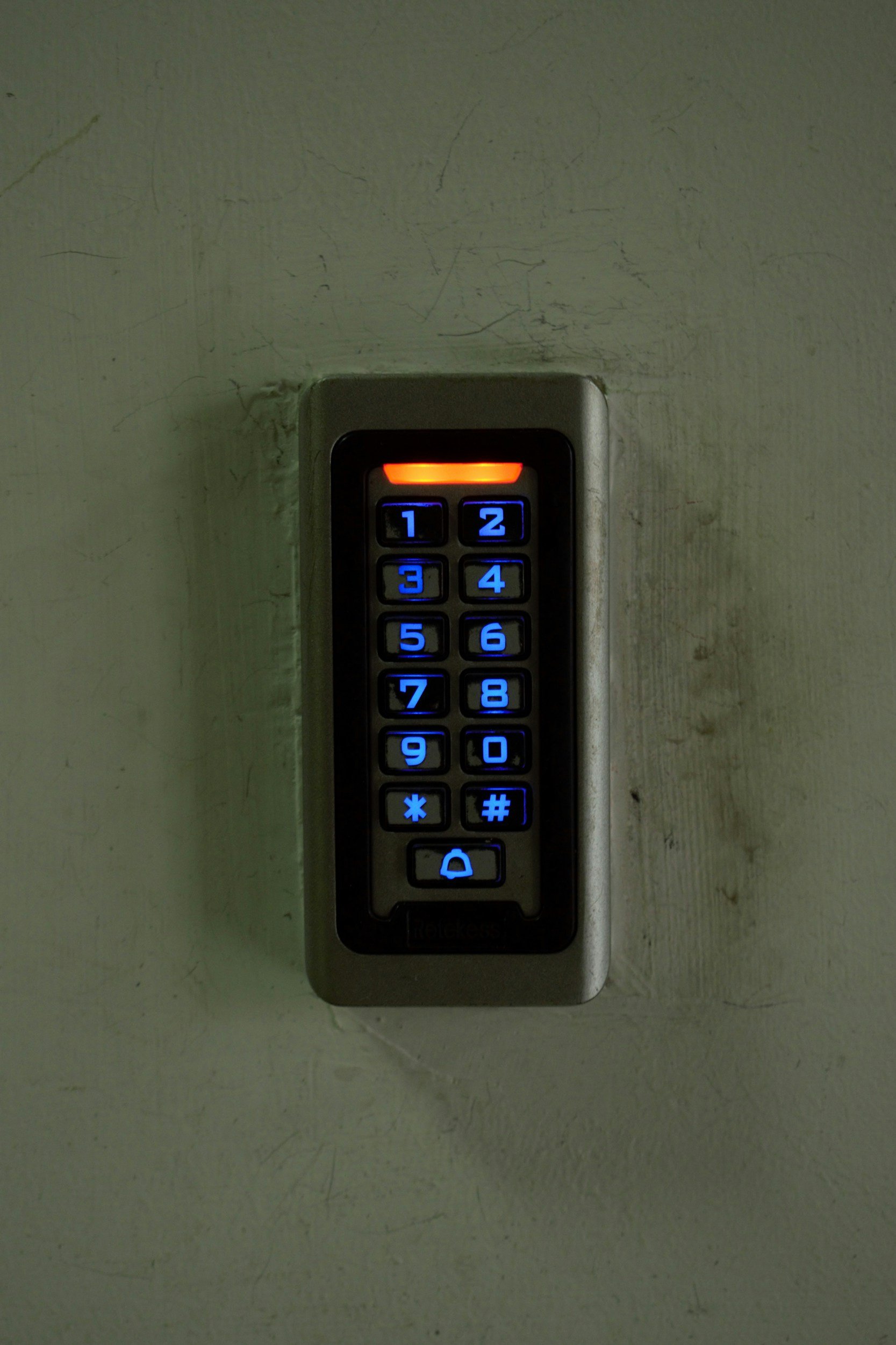 Electronic door keypad with illuminated blue number buttons and a red indicator light at the top mounted on a textured wall.
