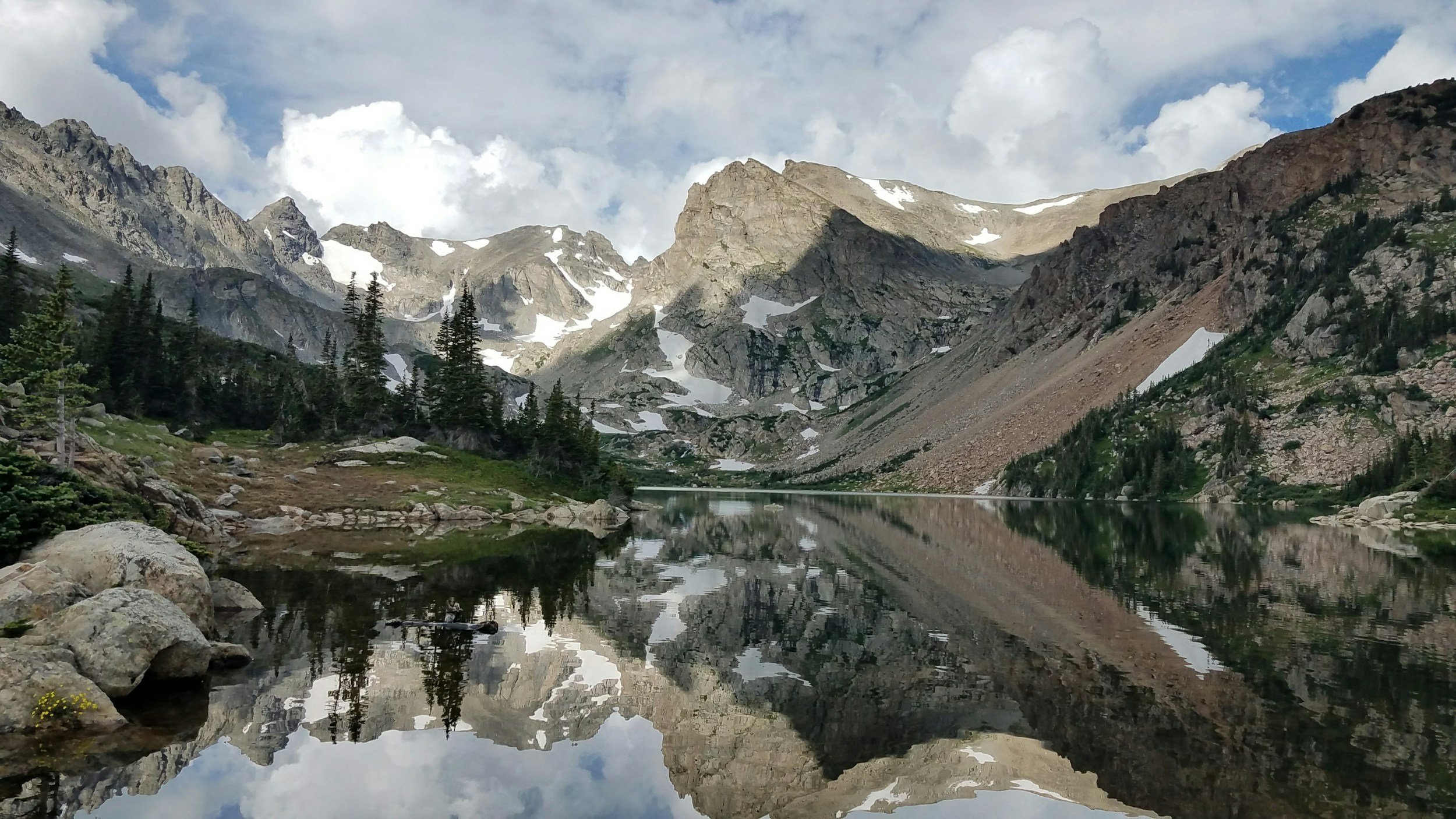 A scenic mountain landscape featuring snow-capped peaks, pine trees, and a calm lake reflecting the mountains and sky.