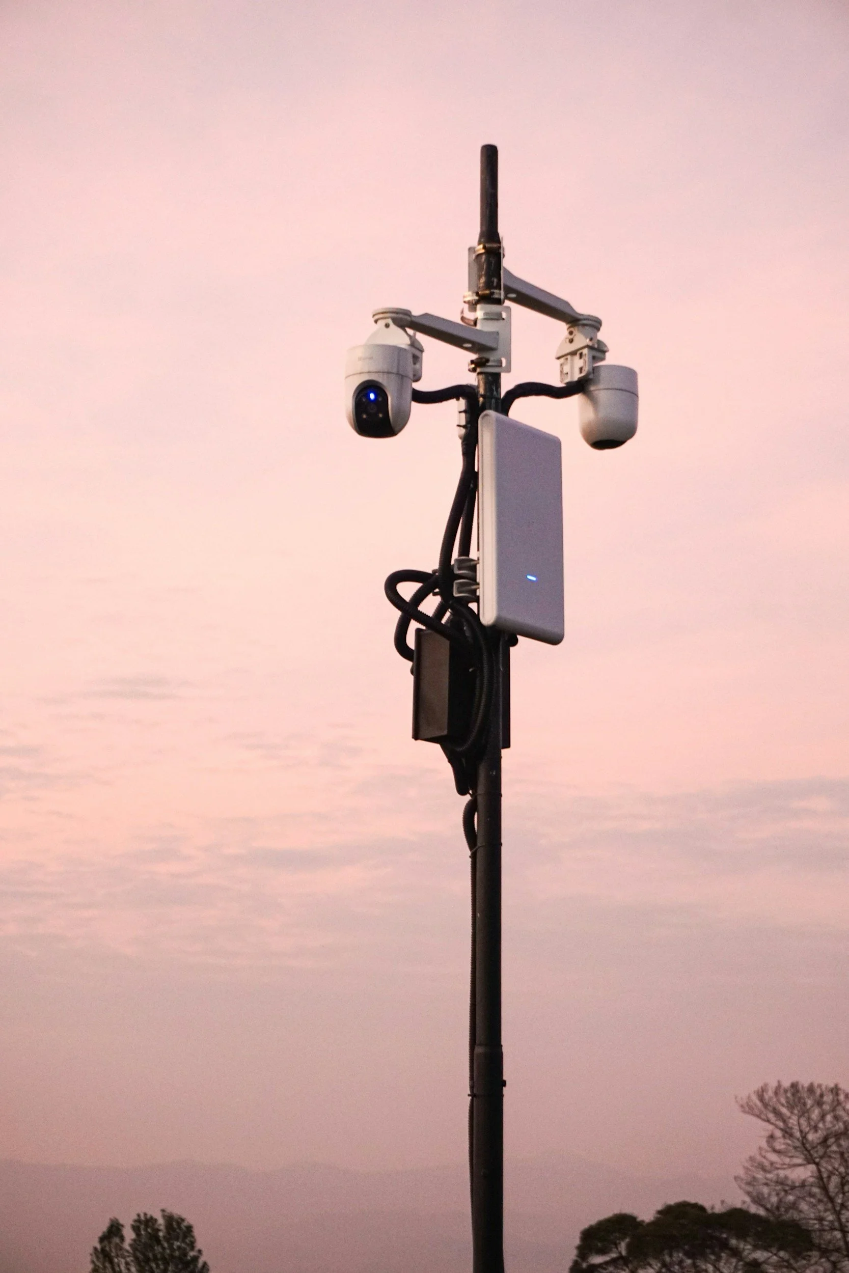 A surveillance camera and sensor equipment mounted on a tall pole against a pastel evening sky with trees in the distance.
