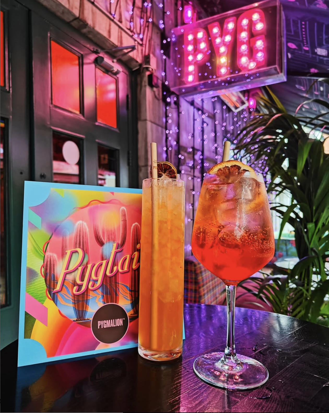 Two colorful cocktails with orange slices on a dark bar table, a menu with tropical design and the word 'Pygmalion', neon signs, purple string lights, and plants in the background.
