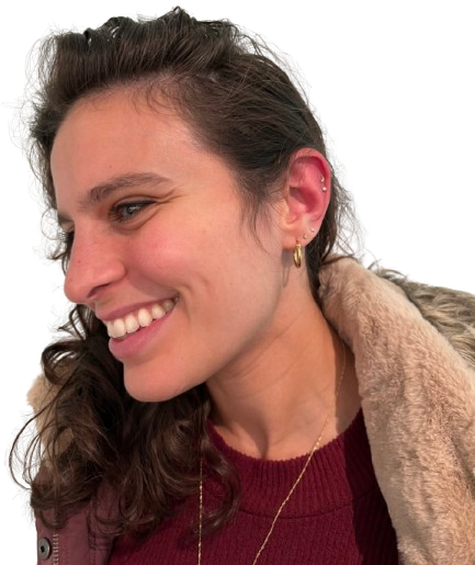 Side profile of a smiling woman with long, wavy brown hair, wearing earrings, a maroon sweater, and a beige coat with a fur collar.