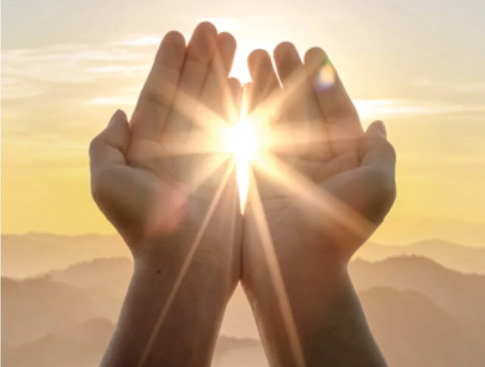 Two hands held together with the sun shining through, creating a starburst effect during sunrise or sunset with mountains in the background.