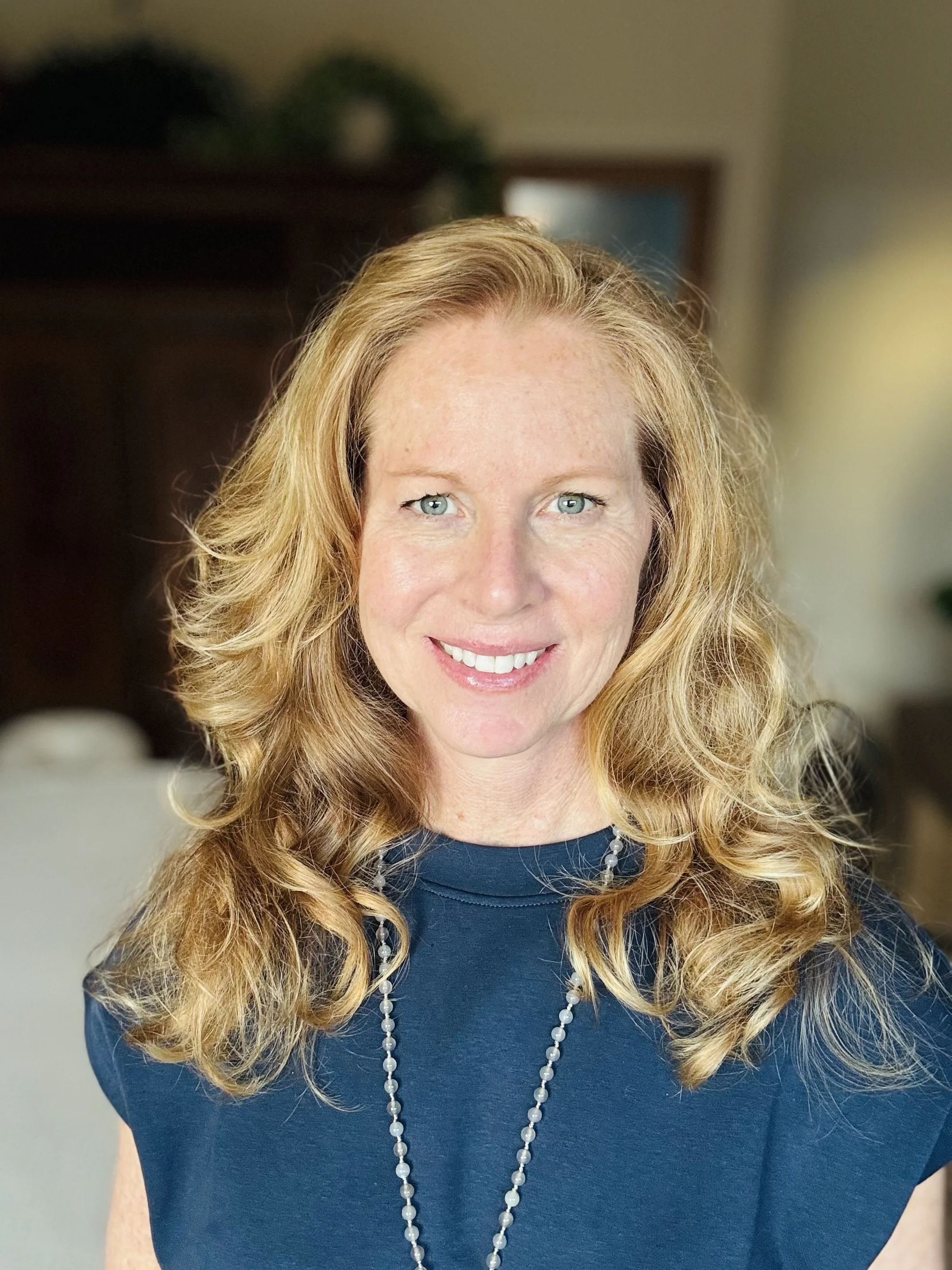 Portrait of a red-haired woman with blue eyes smiling, wearing a navy top and a beaded necklace, indoors with blurred background.