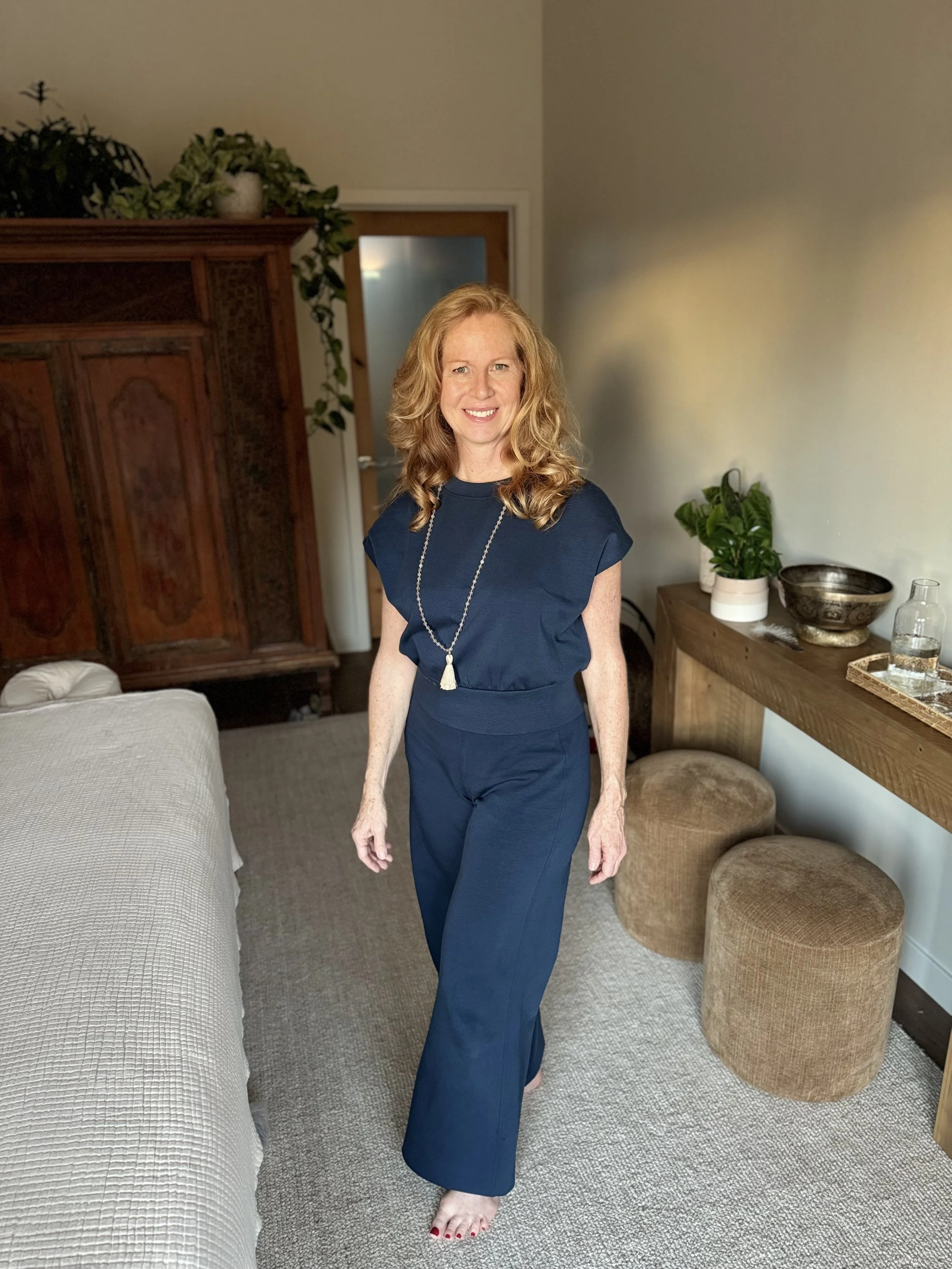 A smiling woman with red hair and fair skin, dressed in a navy blue outfit and necklace, standing barefoot in a cozy room with beige carpet, wooden furniture, and plants.