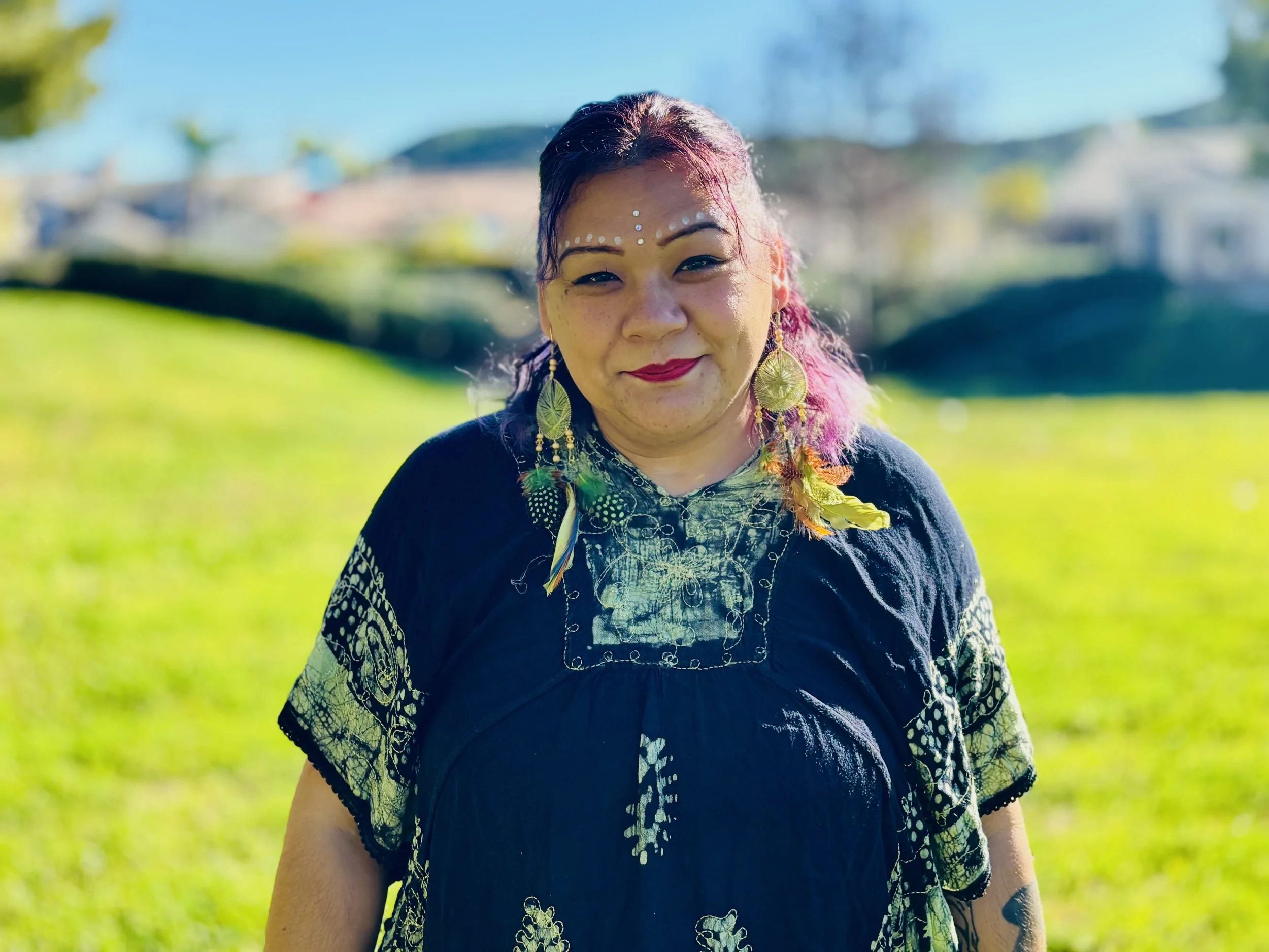 A woman standing outdoors on a sunny day, wearing colorful earrings, a black embroidered top, with makeup and face decorations, in a grassy area with houses in the background.
